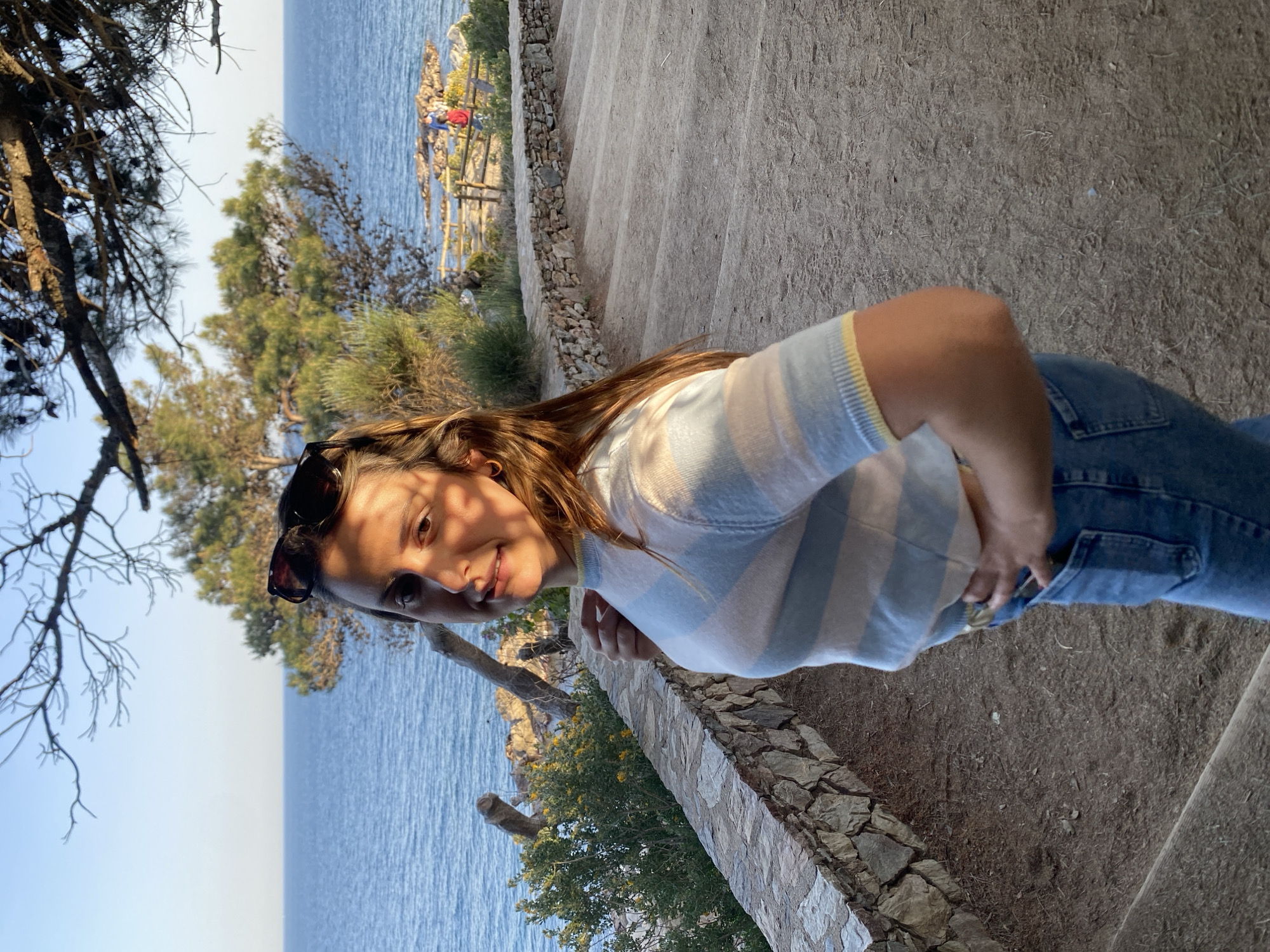 The image is a low-angle, rotated portrait of a woman on a stone staircase near the coast. The woman has long brown hair and is wearing sunglasses, a striped top, and blue jeans. She is smiling slightly at the camera. Behind her, the Mediterranean Sea is visible. To the left, there are trees and vegetation. In the background, a coastal cliff with people and a wooden railing can be seen. The scene is likely located in Calella De Palafrugell, Spain, given the coastal landscape.