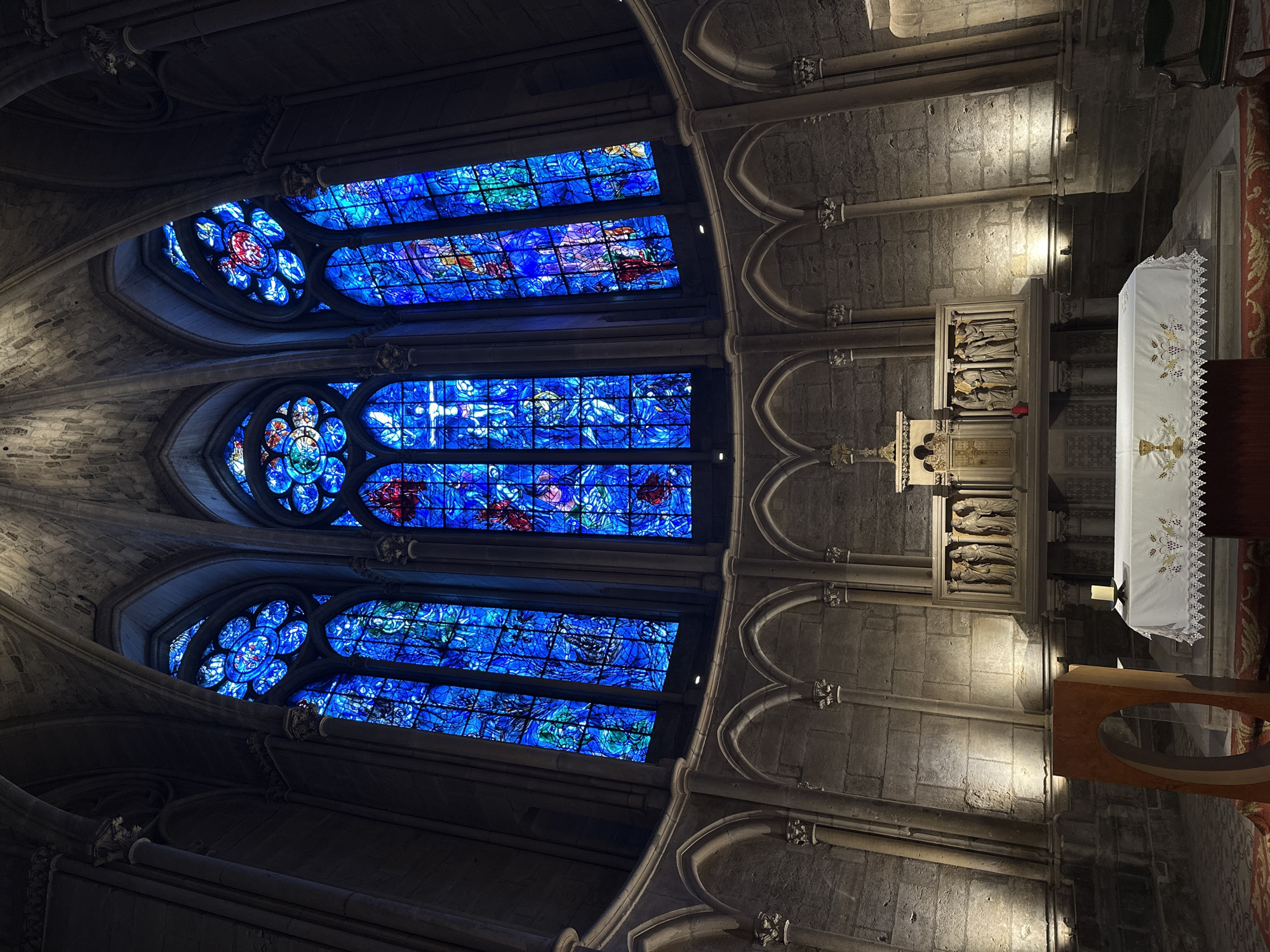 This image captures the interior of the apse of the Reims Cathedral in Reims, France, facing the altar and the magnificent stained-glass windows. The scene is dominated by the vibrant blues and reds of the large, lancet-shaped stained-glass windows above the altar, which are illuminated by natural light. The windows feature intricate religious imagery with figures and abstract patterns.
Below the windows, a stone wall forms the curved apse, with architectural details such as arched niches and carved capitals supporting the vaulted ceiling. The altar is covered with a white cloth, embroidered with gold and purple motifs including a chalice and wheat. A single lit candle sits on a small dark stand to the right of the altar. To the left of the altar, a modern, curvilinear wooden structure, possibly a lectern or a decorative element, stands. In the background, the stone walls of the cathedral are visible, with dramatic spotlights illuminating sections of the stonework. The floor appears to be carpeted in parts with a patterned rug.