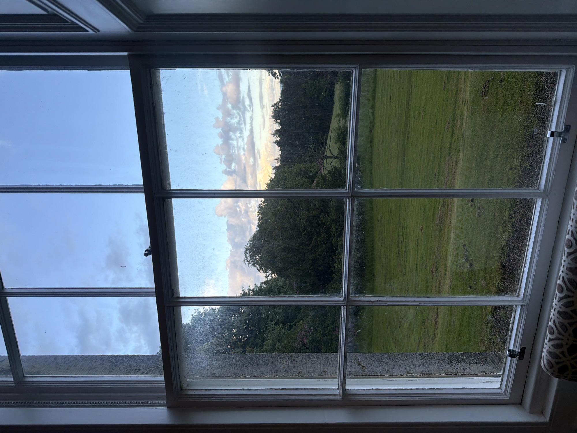 An interior shot looks out through a multi-paned window at a dusky landscape. The window is dirty with streaks and smudges, and the wooden frame is painted white. The sky above is a pale blue with scattered clouds, tinged with pink and orange from the setting sun. Below the sky are dark green trees, some with hints of pink blossoms, and further in the distance, a dense line of darker trees. In the foreground, a neatly mowed green lawn stretches out. A metal latch is visible on the lower section of the window frame. To the left of the window, a stone pillar is visible. In the lower foreground, a patterned upholstered object, possibly a cushion or armrest, is partially visible.