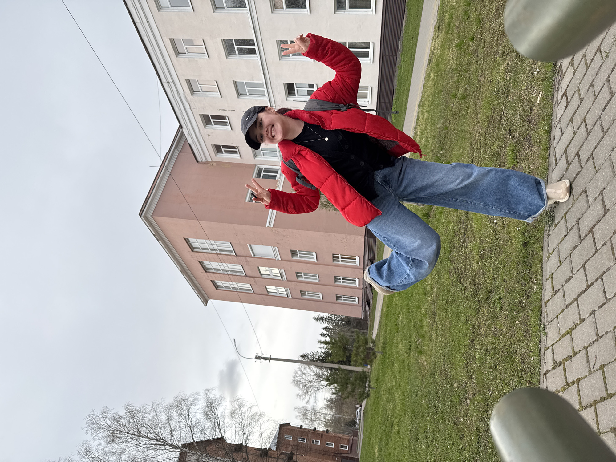 A young woman stands on a paved surface next to a patch of grass. She is wearing a black cap, a red puffer jacket, blue jeans, and light-colored shoes, and she's making the peace sign with both hands. Behind her is a multi-story building with a light pink facade and white-framed windows. Adjacent to this is another building with a beige facade. In the background, trees are visible, along with a street light and brick buildings. The sky is overcast. The location appears to be near a university building in Izhevsk, Russia.