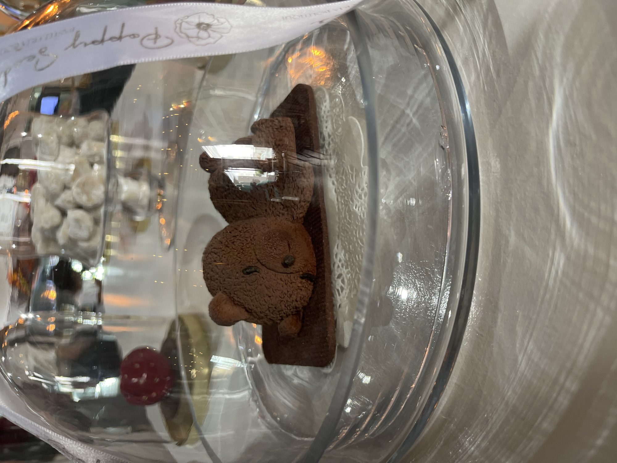The image shows a glass jar containing a chocolate bear-shaped treat, likely a confection or small cake, sitting on a base. The jar is sealed with a ribbon that has the words "Poppy Bea boutique" and a flower logo printed on it. In the background, partially obscured by the glass, are various other treats, including what appears to be a red raspberry-like confection and small, powdery white items. The scene likely takes place in a boutique that sells confectioneries in the city of San Andrés Cholula, Mexico. The photo is taken at an angle, showing reflections and distortions in the glass. The background appears to be a brightly lit area with a plain wall.