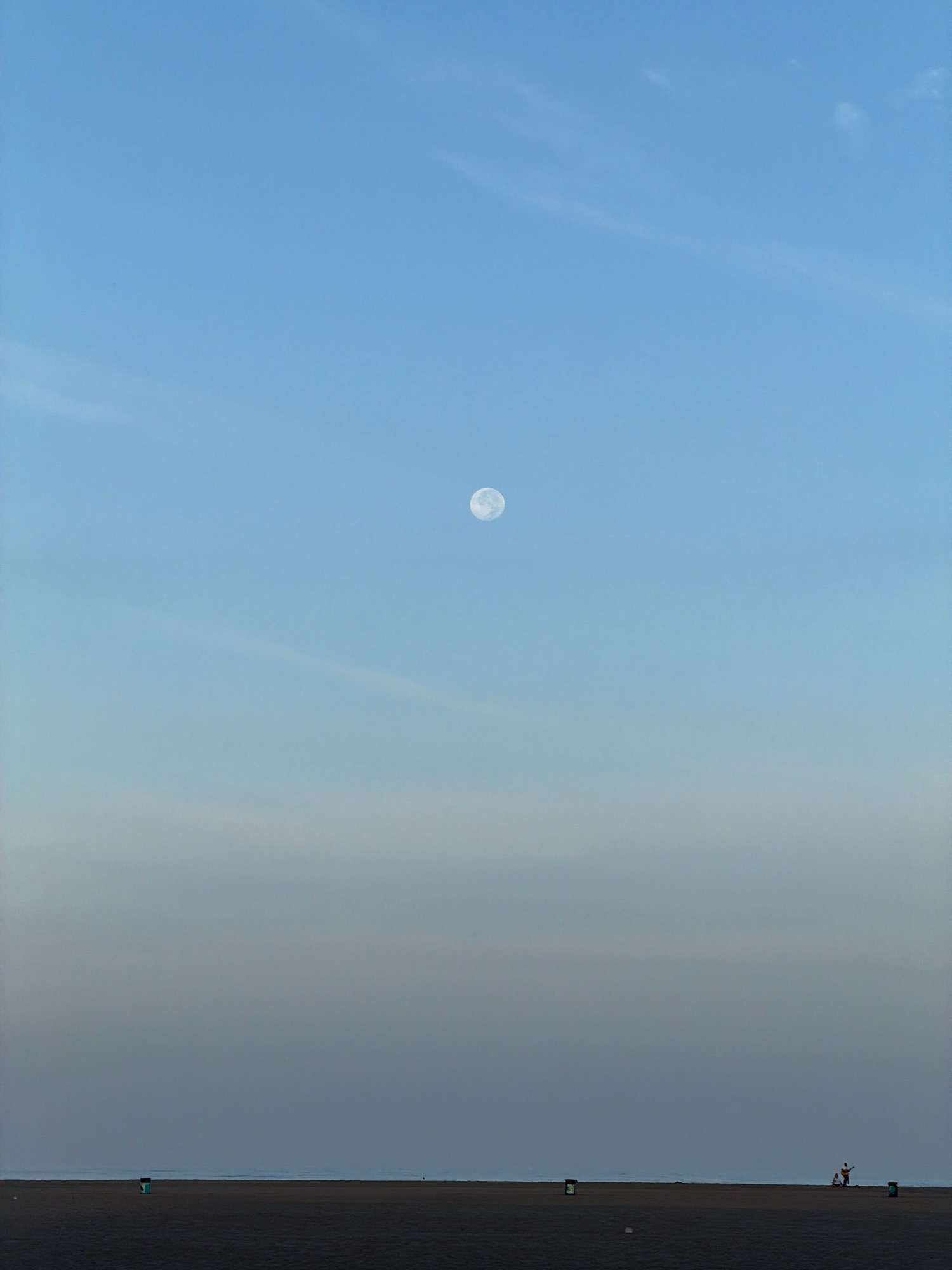 The image shows a wide, flat beach with the ocean and sky dominating the frame. The sky is a gradient of pale blue at the top, fading to a lighter, almost white hue near the horizon. A full moon is visible high in the sky, slightly off-center to the left. The beach itself is a dark, sandy expanse. In the distance, near the horizon, there is a faint line that could be the ocean's edge or distant land. Scattered along the beach are a few small figures. On the left side, near the foreground, a teal and white trash receptacle is visible. Further to the right, a pair of small figures are standing, one of whom appears to be playing a guitar. Another trash receptacle is located near this pair. The overall scene suggests a quiet, serene evening on a Los Angeles beach.