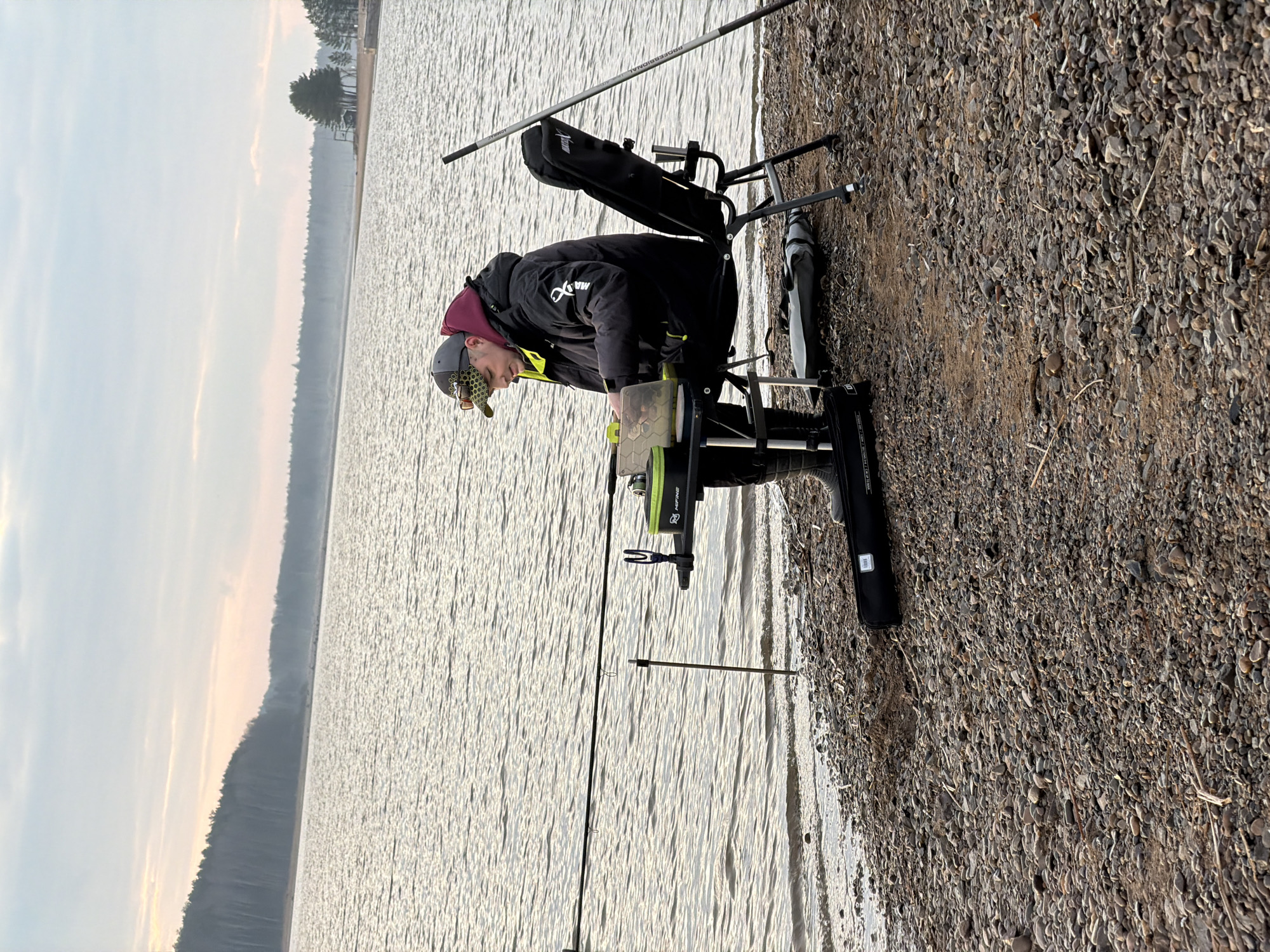 The image depicts a solitary fisherman on the pebbly shore of a large lake in Chaikovsky, Russia, during what appears to be an overcast day. A man, wearing a black jacket with a white "MAX" logo, a maroon hoodie, and a grey cap, is seated on a specialized fishing chair or box. He is leaning forward, intently focused on a light-colored tackle box or container placed on a tray in front of him, likely preparing bait or adjusting his gear.
Several fishing rods are set up around him. One prominent rod, visibly labeled "PROFESSIONAL," extends into the water from a stand, indicating active fishing. The fishing chair setup itself has a "KOFINE" logo visible and features various attachments, including bags and rod holders, suggesting a dedicated and organized angler.
The water surface of the lake is calm with subtle ripples, reflecting the soft, diffused light from the sky. In the background, a distant, dense treeline stretches along the horizon, separating the water from the overcast sky. The shoreline is composed of small stones and gravel. The overall scene conveys a sense of calm and concentration typical of recreational fishing.
