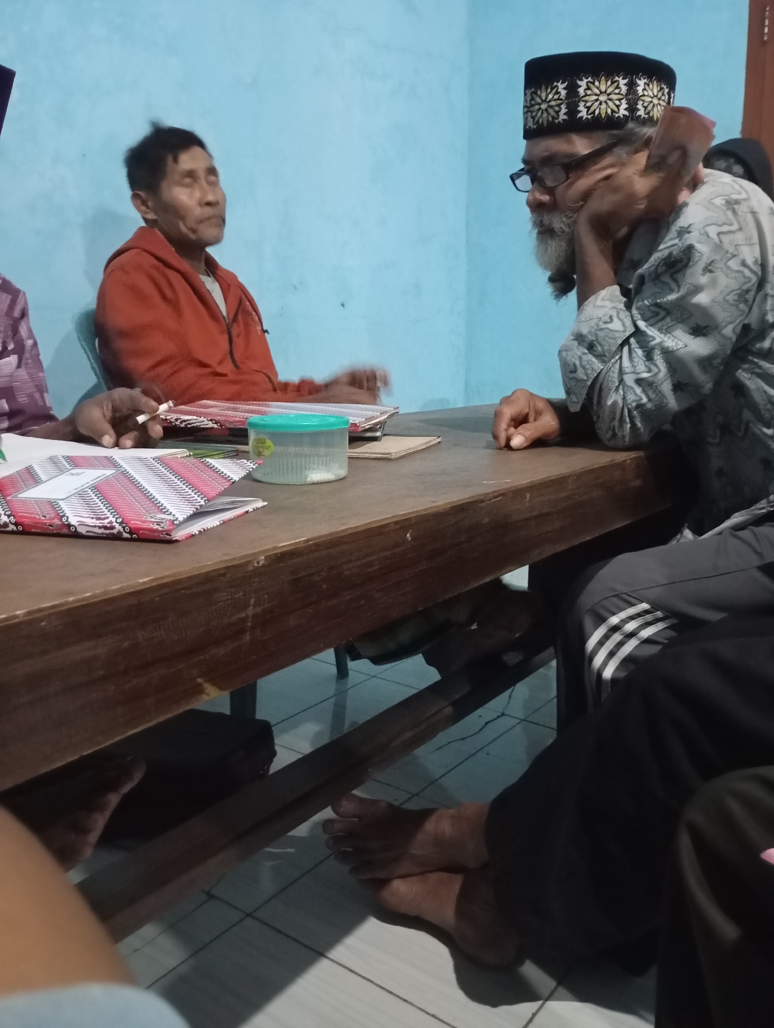 In Ketepung, Indonesia, a group of at least three men are gathered around a wooden table. The man on the right, wearing glasses and a patterned kufi, has a long grey beard and is resting his head on his hand, appearing pensive. He is dressed in a patterned grey shirt. To his left, a man in an orange hooded jacket sits with his eyes closed, possibly smoking. In front of them on the table are several items, including a green lidded container and what appear to be notebooks or binders with patterned covers. The room has light blue walls, and the floor is tiled. The scene suggests a casual indoor gathering.