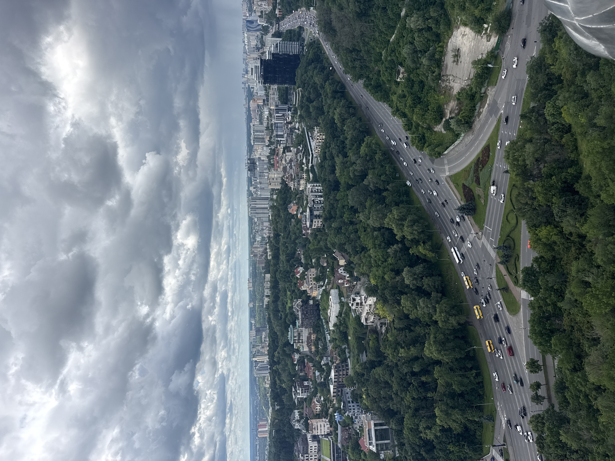 An aerial, wide-angle shot captures a sprawling cityscape in Kyiv, Ukraine, under a heavy, overcast sky. The scene is dominated by a dense forest of green trees that separates the foreground from the background. A multi-lane highway, teeming with cars and a few yellow buses, cuts through the landscape, curving its way towards a distant cluster of high-rise buildings. Interspersed within the greenery are various residential buildings, from multi-story apartment complexes to more modest houses with red-tiled roofs. The sky itself is dramatic, filled with thick, gray clouds that hint at impending rain. The overall impression is one of urban development encroaching upon a natural, verdant environment.