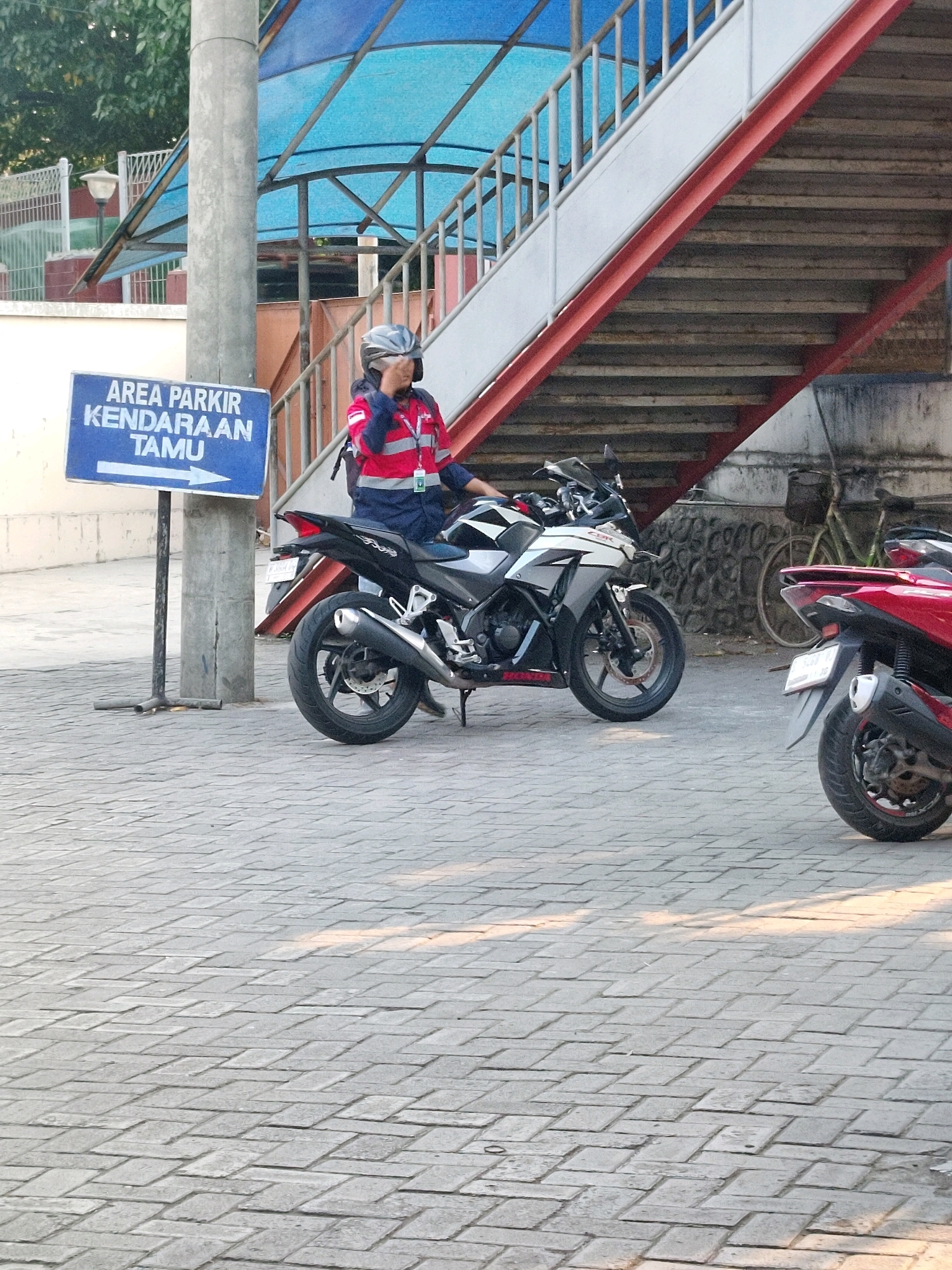 A person wearing a helmet and a red and blue uniform is sitting on a black and white motorcycle. The person appears to be adjusting their helmet. The motorcycle is parked on a paved area made of interlocking bricks. A blue sign with white text that reads "AREA PARKIR KENDARAAN TAMU" and an arrow pointing to the right is visible to the left of the motorcycle. Behind the motorcycle, there is a concrete staircase with red railings. Further in the background, a bicycle is visible, leaning against a stone wall. To the right of the black and white motorcycle, the rear of a red scooter is partially visible. The scene is outdoors, with sunlight casting shadows on the ground. The location is identified as Kramat Temenggung, Indonesia.
