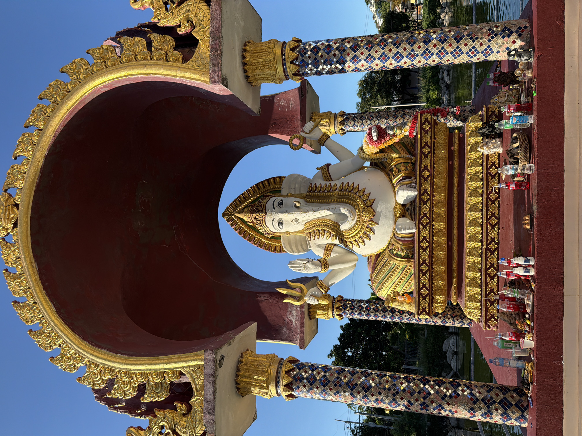 A Ganesha statue is situated in an alcove at Baan Plai Laem temple in Koh Samui, Thailand. The statue is white with gold ornamentation and wears a crown. It is seated on a golden throne, with offerings of bottles and food placed on a red platform in front of it. The alcove is framed by ornate golden arches and supported by two pillars decorated with colorful mosaic tiles. The background shows a blue sky and lush greenery, with a body of water to the right of the statue.