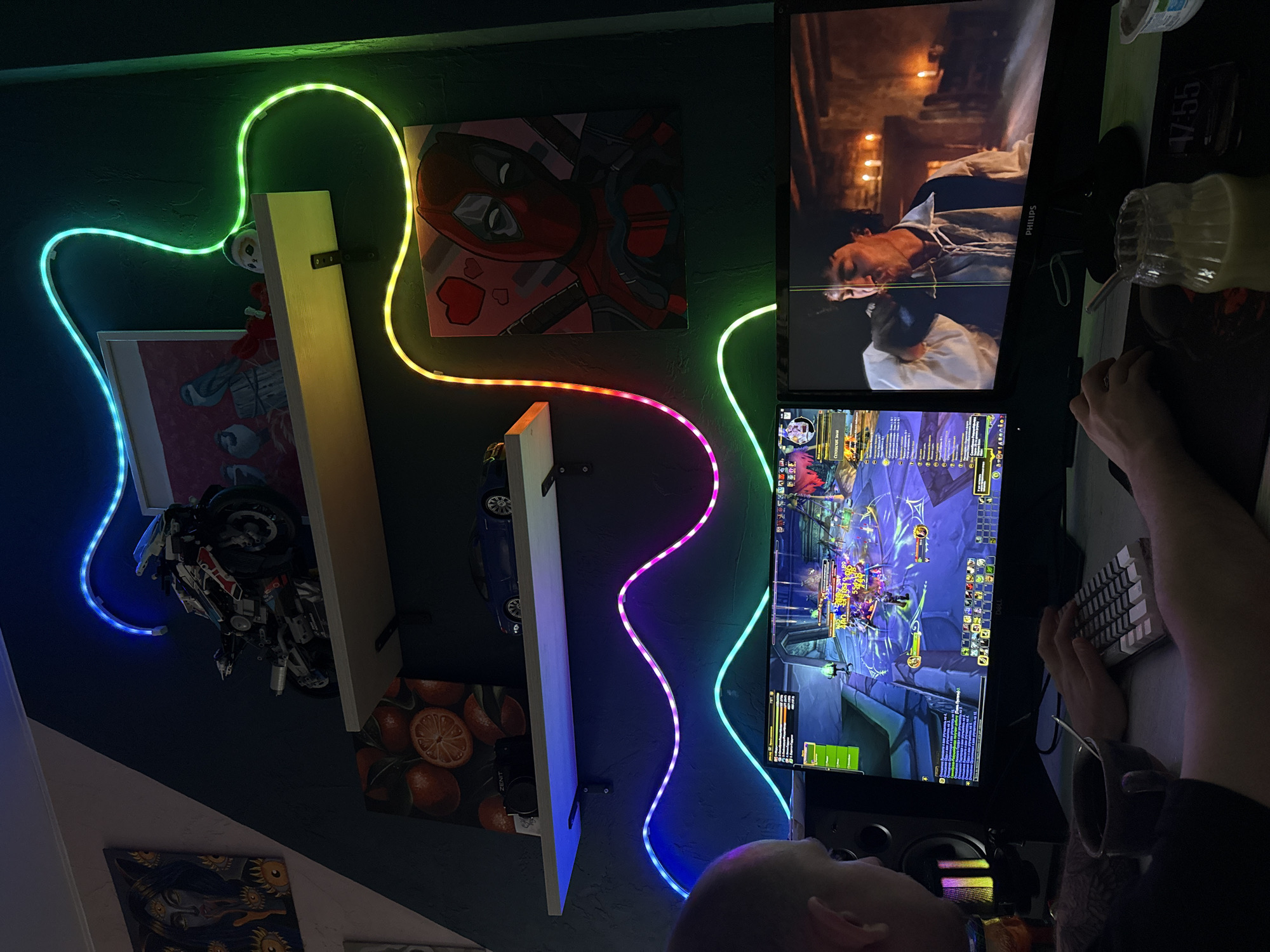 A person is playing a video game on a dual-monitor setup in a dimly lit room. The upper monitor displays a movie scene, while the lower monitor shows a game interface with a fantasy-style world. The person is using a small, white keyboard and their hands are visible on the keyboard. The camera angle is a high-angle, looking down and slightly to the side of the person. There is a rainbow-colored LED light strip illuminating the wall and shelves. The shelves hold various items including artwork, a model motorcycle, a model car, and a drawing of oranges. It cannot be determined whether or not this scene is located in Rostov-on-Don, Russia.