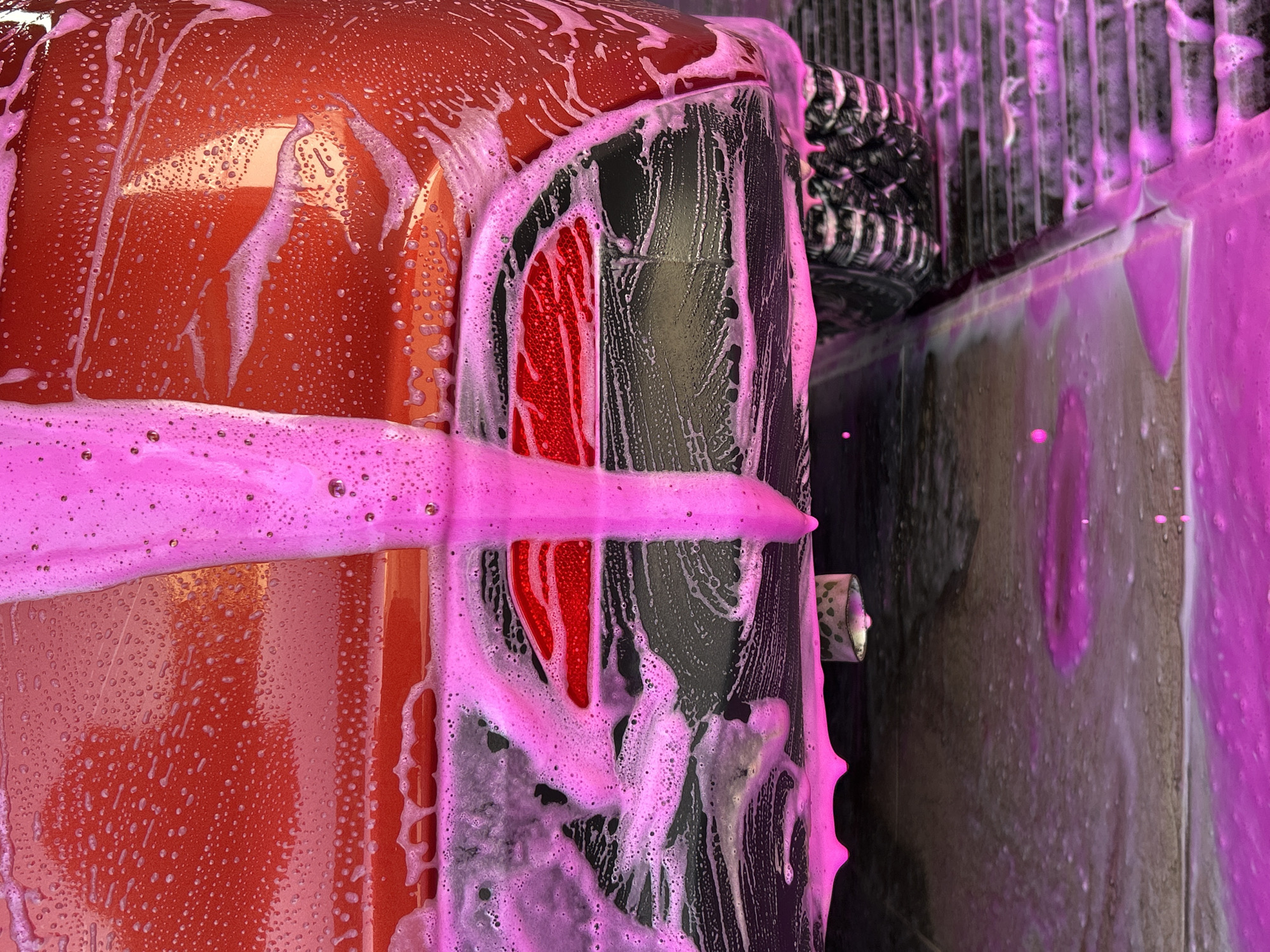 A close-up, side-view shot shows a section of a vehicle undergoing a wash. The vehicle's body is predominantly orange, and a portion of the rear light, which is red, is visible. Pink soap suds heavily coat the car's surface, dripping and clinging to its contours. Adjacent to the car, a wall-like structure with vertical lines is also covered in the same pink suds. To the upper right, a black, textured object appears to be part of the carwash equipment. The exact location is unidentifiable from this image alone, and I am unable to verify that this picture was taken in the city of Chaikovsky, Russia.