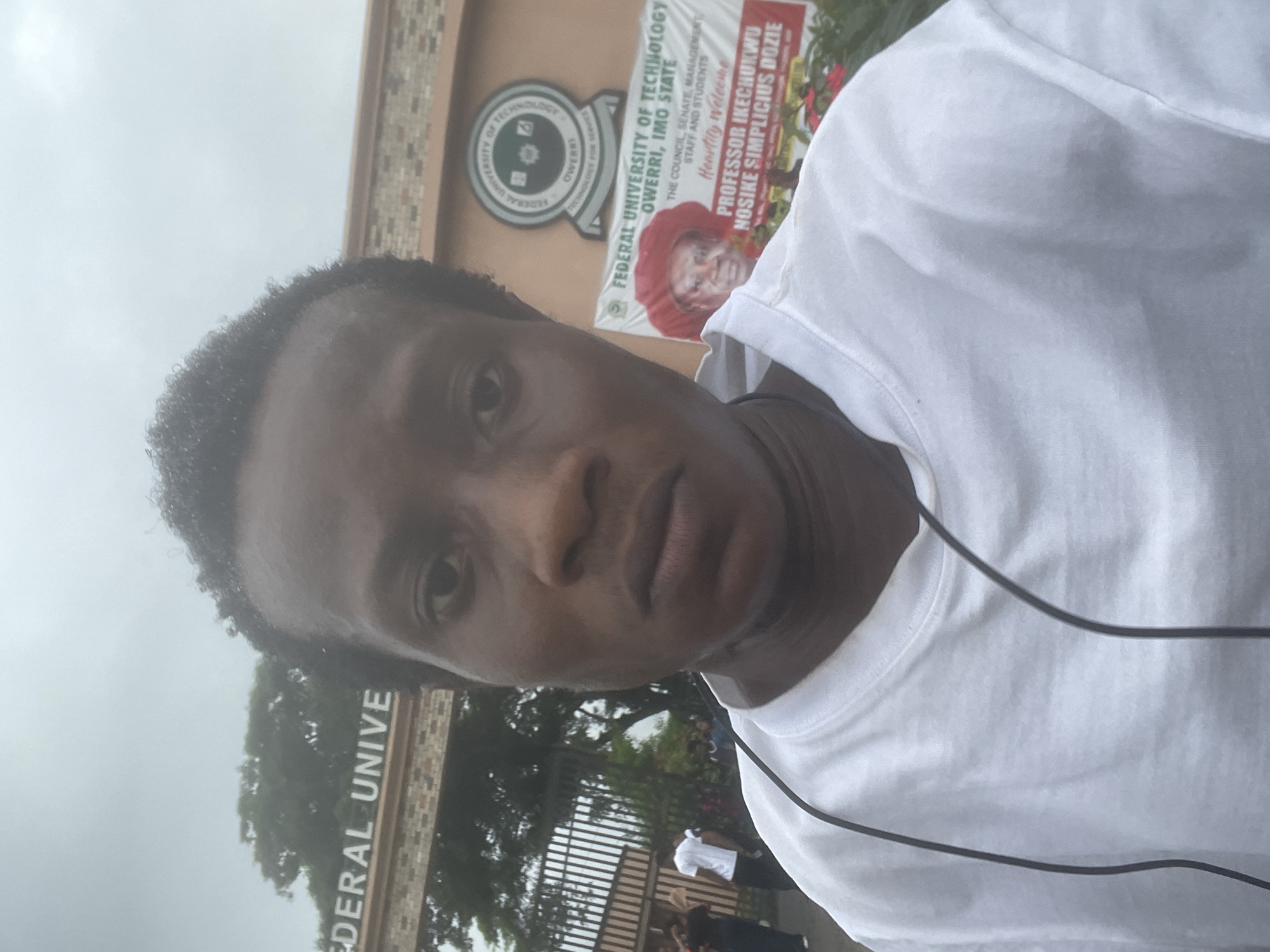 A young Black male, appearing to be in his late teens or early twenties, takes a selfie outside a university. He is wearing a plain white t-shirt and has dark, short, curly hair. A dark wire, possibly from headphones or a microphone, is visible hanging around his neck and across his shirt.
Behind him, the entrance to the Federal University of Technology, Owerri (FUTO) is visible. The university's sign is above an archway, with the words "FEDERAL UNIVE" clearly legible. A large banner is also displayed, announcing "FEDERAL UNIVERSITY OF TECHNOLOGY OWERRI, IMO STATE" and bearing a portrait of a man in a red cap, identified as "PROFESSOR IKECHUKWU NOSIKE SIMPLICIOUS DOZIE". The background suggests an outdoor, possibly overcast day, with trees and other people visible in the distance near the university gates. The overall scene indicates the individual is on the grounds of the FUTO campus in Ihiagwa, Nigeria.