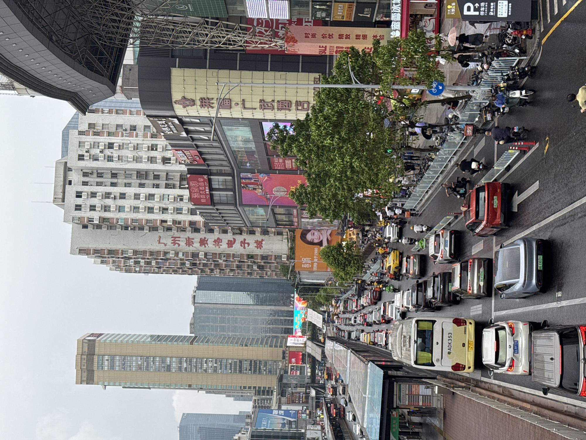 The image shows an elevated view of a congested street in Tianhe District, China. The street is densely packed with cars, buses, and electric scooters. Several pedestrians are seen near the roadside. Towering buildings with commercial signs and advertisements line the street. The sky is overcast.