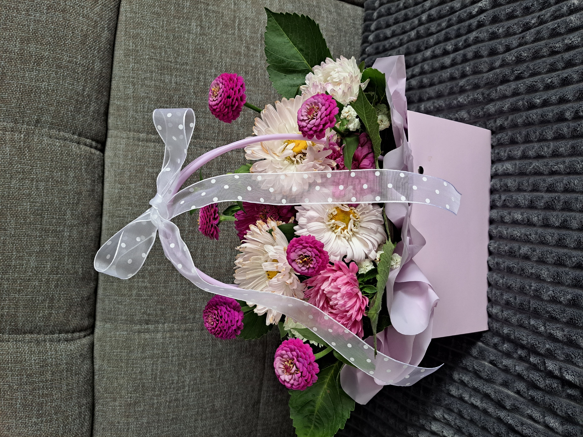 This is a photograph of a pink gift box filled with a floral arrangement. The flowers are a mix of pink and white asters, with some deep pink zinnias. The box is decorated with a white ribbon with pink polka dots that forms a bow at the top. The box is resting on a dark grey, textured surface, possibly a couch or blanket. Behind the box is a dark grey upholstered surface, likely the back of a couch. There are no people or animals visible in the image. The lighting suggests it is daytime, with soft illumination. The image does not provide any specific location information within the city of Chernivtsi, Russia.