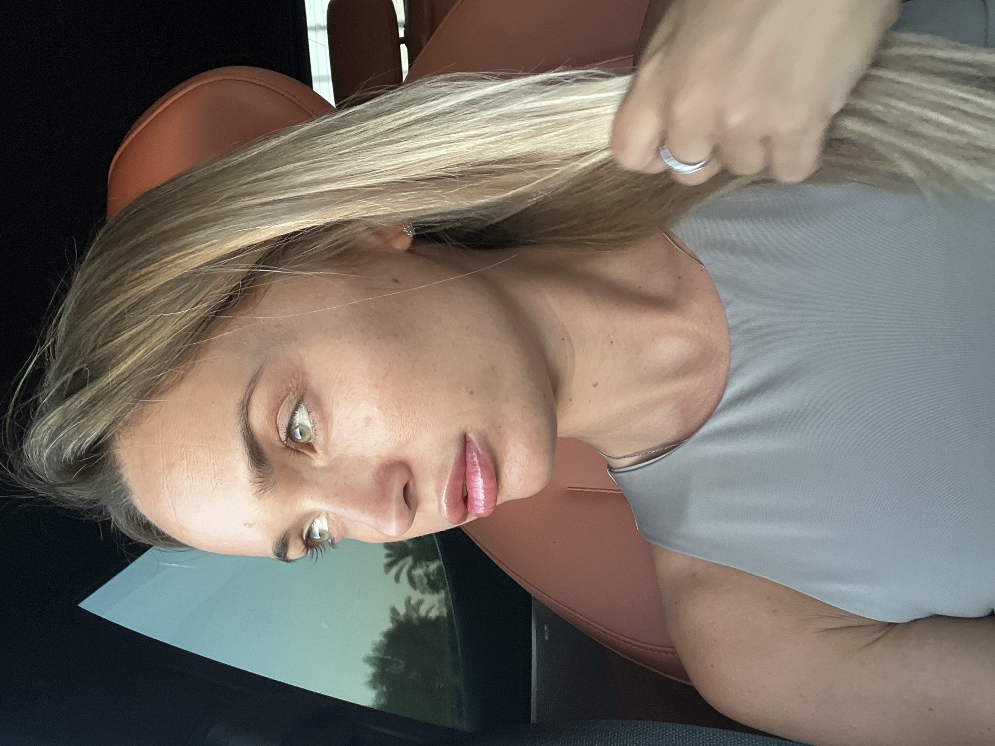 A medium shot captures a Caucasian woman with blonde hair and green eyes sitting in a car. She is looking to her left, out of the car window, with her mouth slightly open. Her right hand is touching her hair, and she is wearing a grey sleeveless top, a silver necklace, a diamond stud earring, and a ring on her right hand. The interior of the car features brown leather seats. Outside the window, palm trees are visible against a clear sky. The scene appears to be in Sharm El Sheikh, Egypt, possibly during daylight hours.