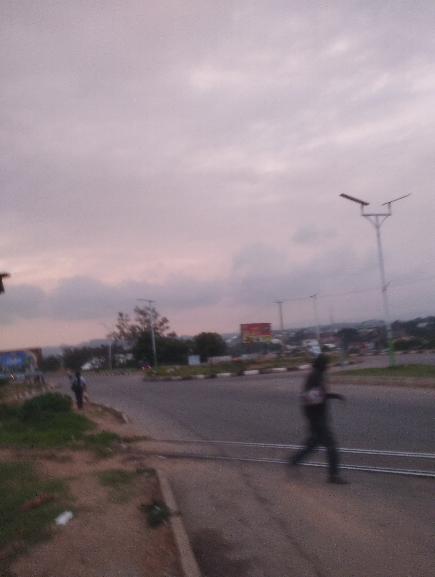 The image captures a street scene in Jos, Nigeria, under a cloudy sky at dusk. In the foreground, a person is walking across a road with double white lines, their form blurred by motion. To the left, a grassy embankment slopes down towards the road, with some litter visible. Further back, a multi-lane road stretches into the distance, lined with streetlights and bordered by a roundabout with decorative planters. Beyond the road, a cityscape unfolds with buildings and trees, and a large billboard with red and yellow signage stands out. The overall impression is one of a typical urban environment during the transition from day to night.