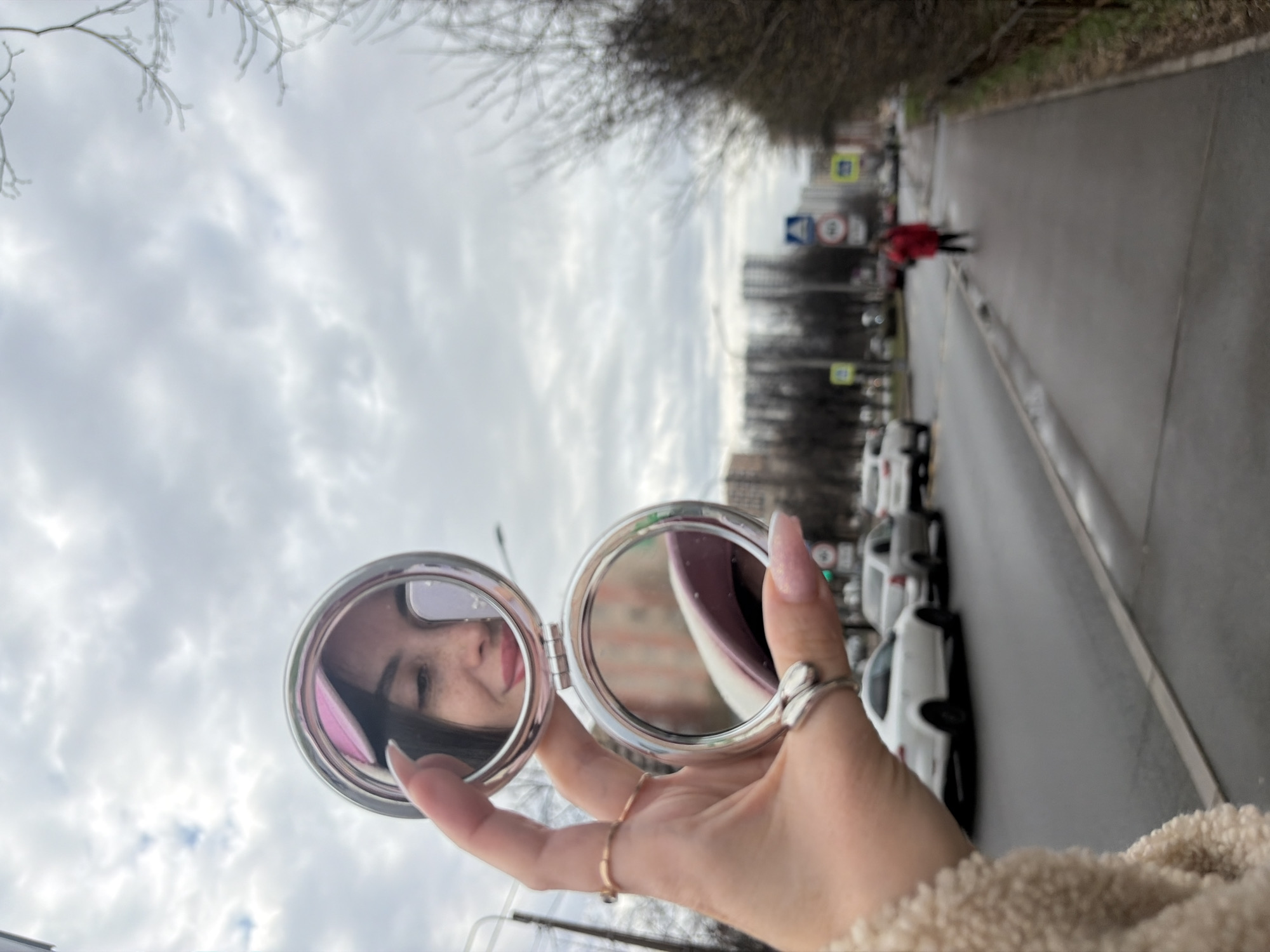 A woman holds a double-sided compact mirror reflecting her face in front of an urban scene. Her hand, adorned with rings and manicured nails, grips the mirror.
In the background, a wet asphalt road stretches away into the distance. Several parked cars are visible along the road's edge. Trees line the opposite side of the road, and a cloudy sky fills the upper portion of the frame. There is a person walking away on the sidewalk in a red jacket. Road signs are also visible in the background. Based on the architecture and street signs, it is possible that this image was taken in the city of Saint Petersburg, Russia.