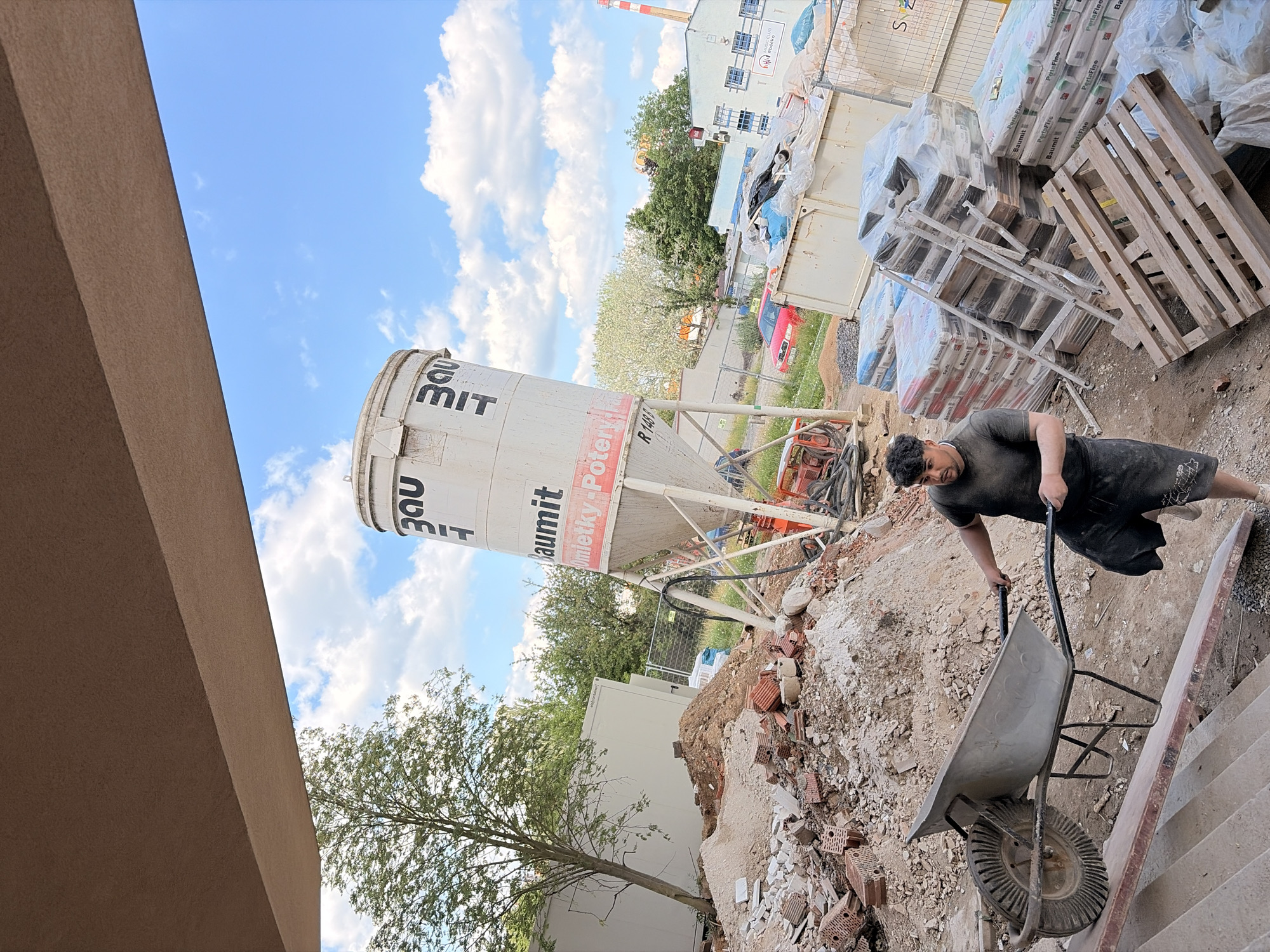 This media file captures a construction site scene in Příbram, Czechia, under a bright, cloudy sky, suggesting daytime. The central focus is a man pushing a wheelbarrow up a set of concrete stairs. He is wearing a dark t-shirt and shorts, and appears to be working on the site.
Dominating the background is a large, white cement silo with prominent black lettering that includes "3DU MIT" and "Baumit Omietky-Potery". Beside the silo, stacks of building materials, primarily bags of what appears to be cement or plaster, are wrapped in plastic. These bags display text such as "Baumit" and "PerlaFine". Wooden pallets are also visible, supporting some of the stacked materials.
In the mid-ground, there's a glimpse of a red car, and a chain-link fence separates the construction area from a green, treed background. Further in the distance, a building with multiple windows and a red and white striped smokestack is visible, along with other construction containers. The ground is covered in dirt, rubble, and scattered bricks, typical of an active construction environment. The overall impression is one of ongoing construction work.