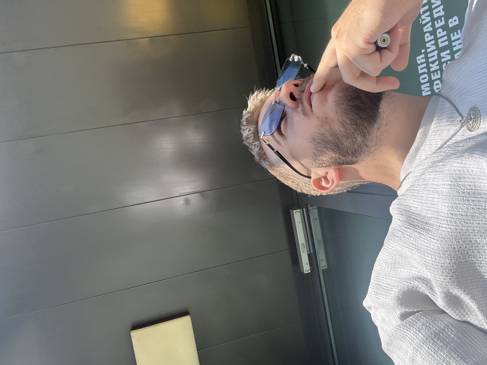 The image depicts a man in Sofia, Bulgaria, wearing sunglasses and a white shirt. He is standing next to a dark gray paneled wall with door hardware visible, and an illuminated panel. The man has short blond hair and a beard. In his hand, he holds an electronic cigarette device up to his mouth. There is text visible on a sign to the right of the man, indicating the location is likely in a public area.