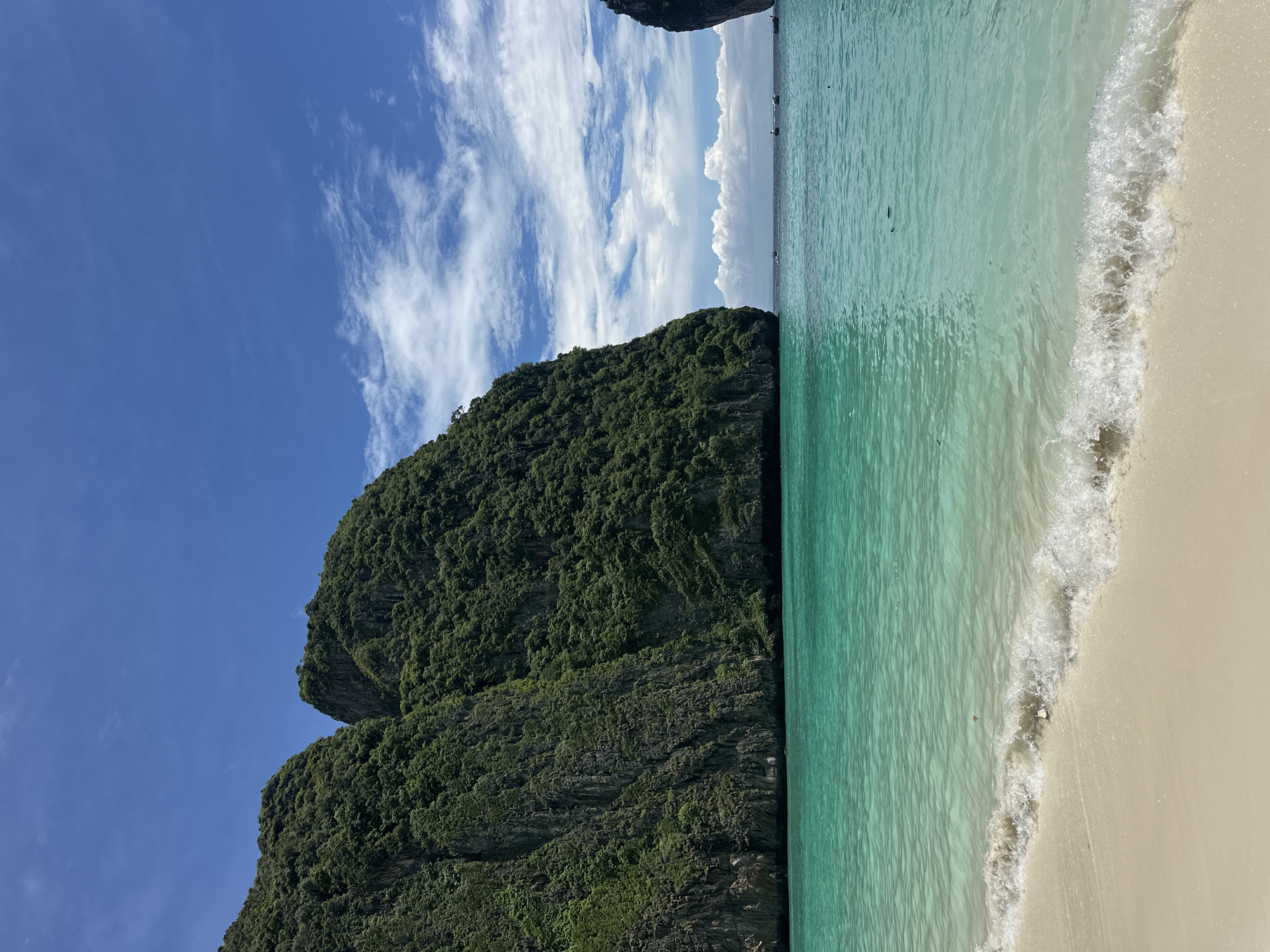 The image depicts Maya Bay, located in the Hat Noppharat Thara-Mu Ko Phi Phi National Park, near Ao Nang, Thailand. The scene is a tropical beach with a shoreline of light-colored sand. Turquoise-colored water with gentle waves laps at the shore. To the left and in the background, a large, verdant limestone cliff rises from the sea, covered in dense green vegetation. The sky above is a bright blue, with scattered white clouds. In the distance, several small boats are visible on the water, and another limestone formation can be seen to the right. There are no people visible in the frame.