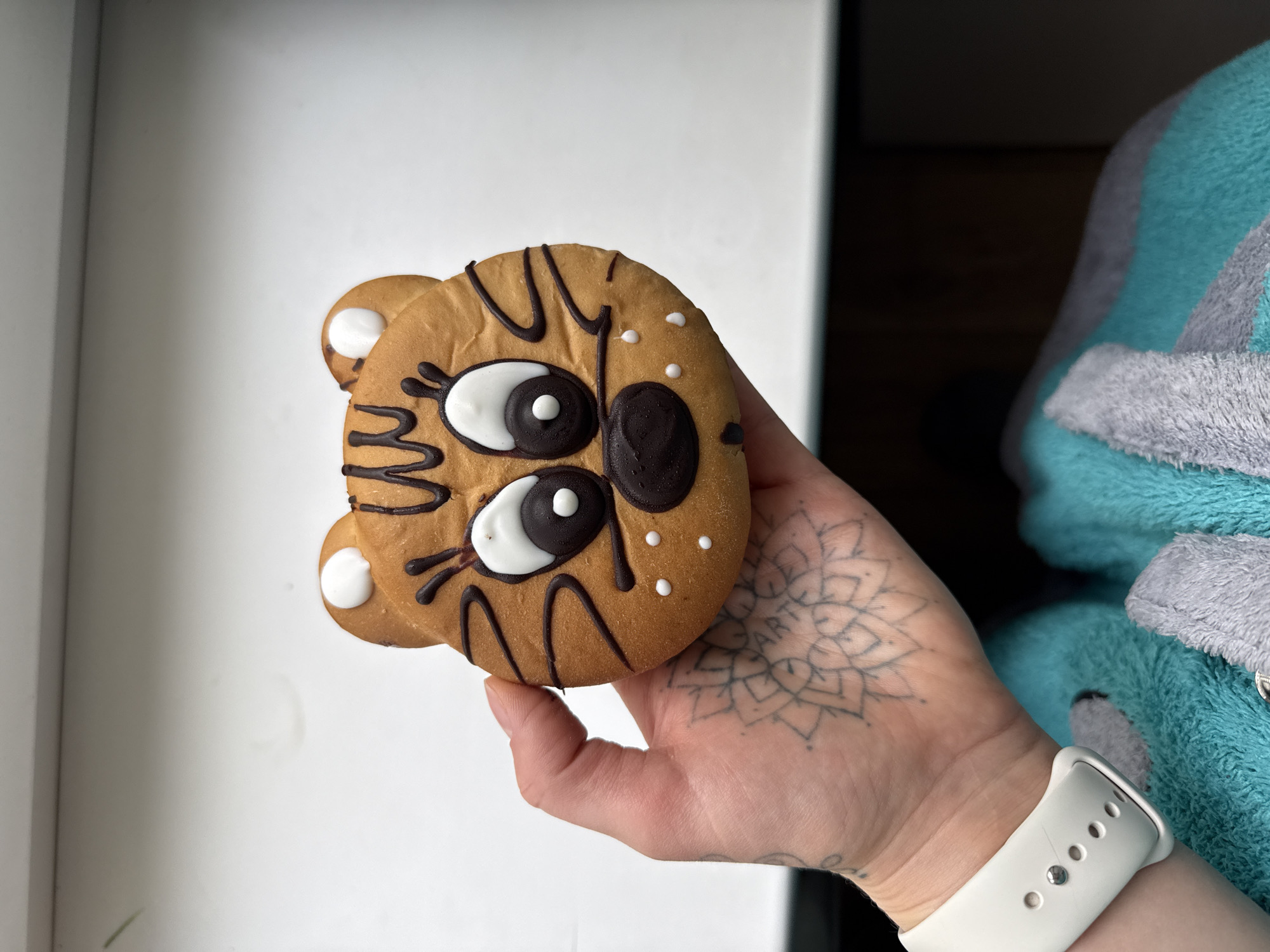 A hand with a tattoo is holding a cookie shaped like a tiger's face. The cookie is decorated with brown and white icing. The hand is wearing a white Apple Watch band. The background is a white wall and a blurry blue and gray object. The location is not specified in the prompt.