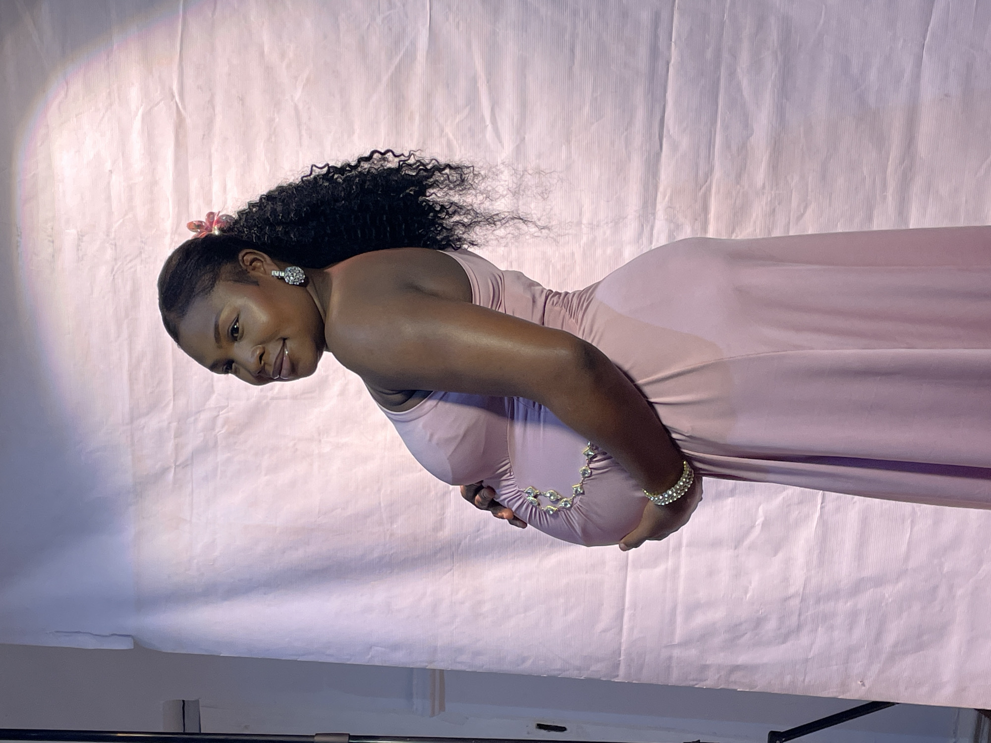 The image shows a pregnant woman in a light pink dress standing against a pink backdrop. She is holding her stomach with both hands, and a jeweled accessory adorns her midsection. The woman has dark, curly hair, and is wearing earrings and a flower in her hair. The lighting in the scene appears to be artificial, with a bright spot visible on the backdrop. Based on the prompt, this photograph was taken in Barakin Akawo, Nigeria, though there are no elements in the picture that would verify this. There is a visible stand beneath the backdrop that does not occupy the main view of the image.