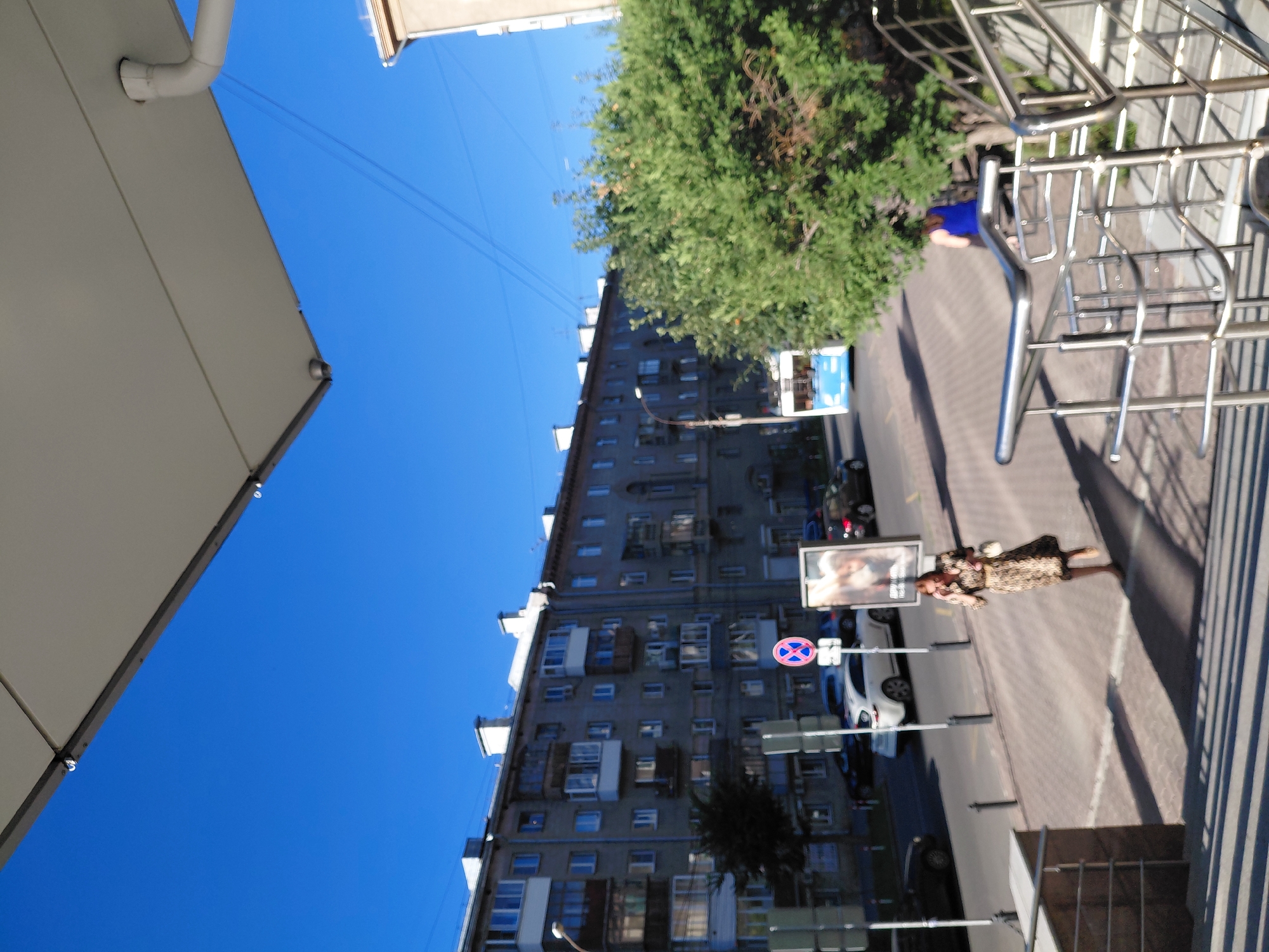 The image depicts a street scene in Novosibirsk, Russia, under a clear blue sky. In the foreground, a metal staircase railing is visible, suggesting an elevated viewpoint or entrance. The middle ground shows a woman in a patterned dress walking away from the camera, holding a white purse, and appearing to be on her phone. She is on a paved walkway beside a street. On the street, a white car and a blue bus are visible, along with other parked cars. Behind the street and walkway, a multi-story apartment building with balconies lines the road. A tall green tree is also present. The overall atmosphere is that of a sunny day in an urban environment.