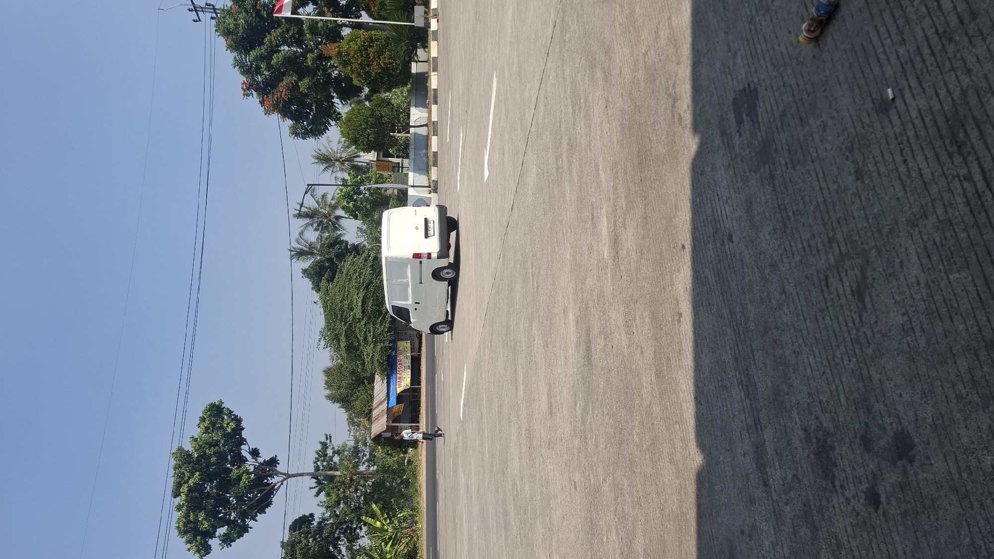 A white panel van is parked in a paved lot. A person in a light-colored shirt and dark pants walks away from the van on the left side of the image. In the background, there is a small building with a blue and yellow awning. Trees, including palm trees, are visible behind the building and along the horizon. The sky is clear and blue. The ground of the lot has painted white directional arrows. A shadow is cast on the lower portion of the image. A small object, possibly a cigarette butt, and some colorful cylindrical objects are on the ground in the lower right.