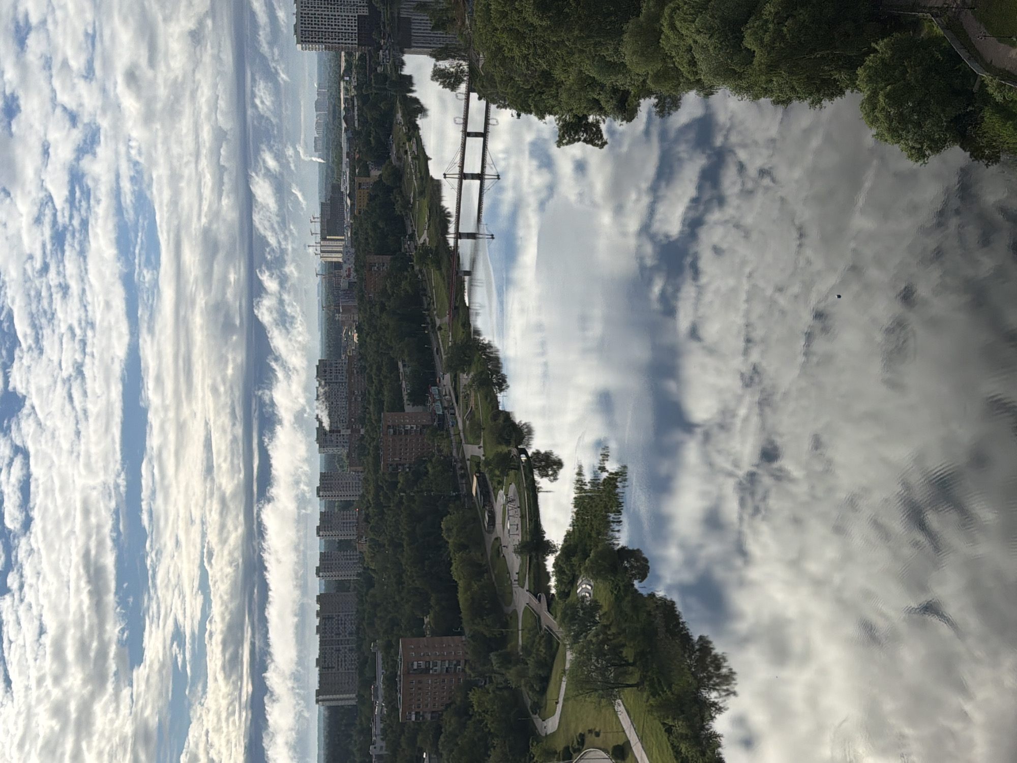 The image shows a landscape with a river running through an urban area. The river reflects the sky, which is filled with long, stretched clouds. Buildings are visible along the riverbank, with trees and green spaces interspersed. A bridge crosses the river. The perspective is from an elevated position, looking down on the scene. The location appears to be a city, although the specific city and country are not identifiable from the image.