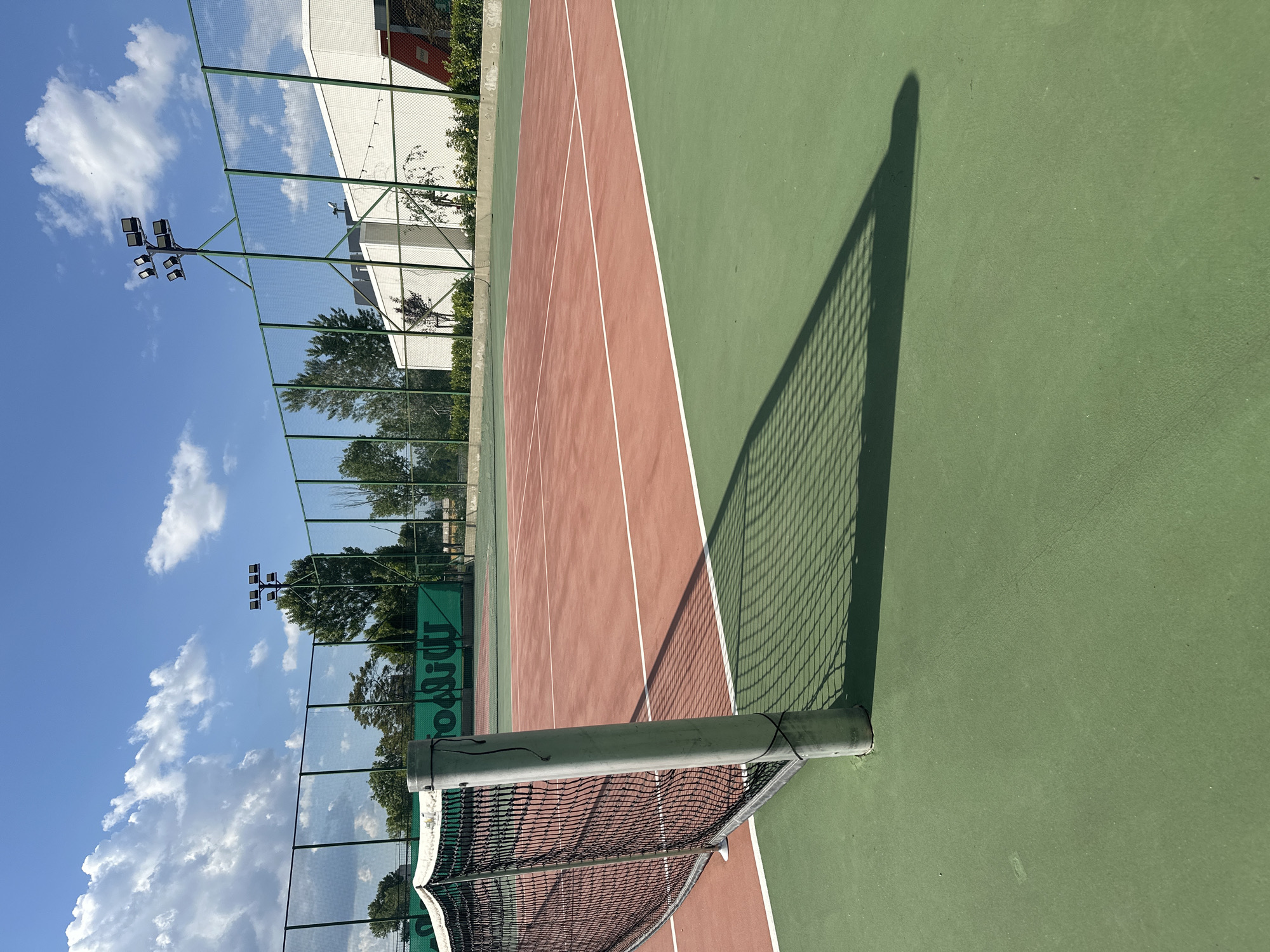 The image shows an outdoor tennis court with a green and reddish-brown surface. A tennis net divides the court. The shadow of the net and the supporting pole is cast on the green part of the court. The background features a green fence, trees, a white building, light poles, and a blue sky with clouds. The image was taken during daylight, likely in Sofia, Bulgaria.