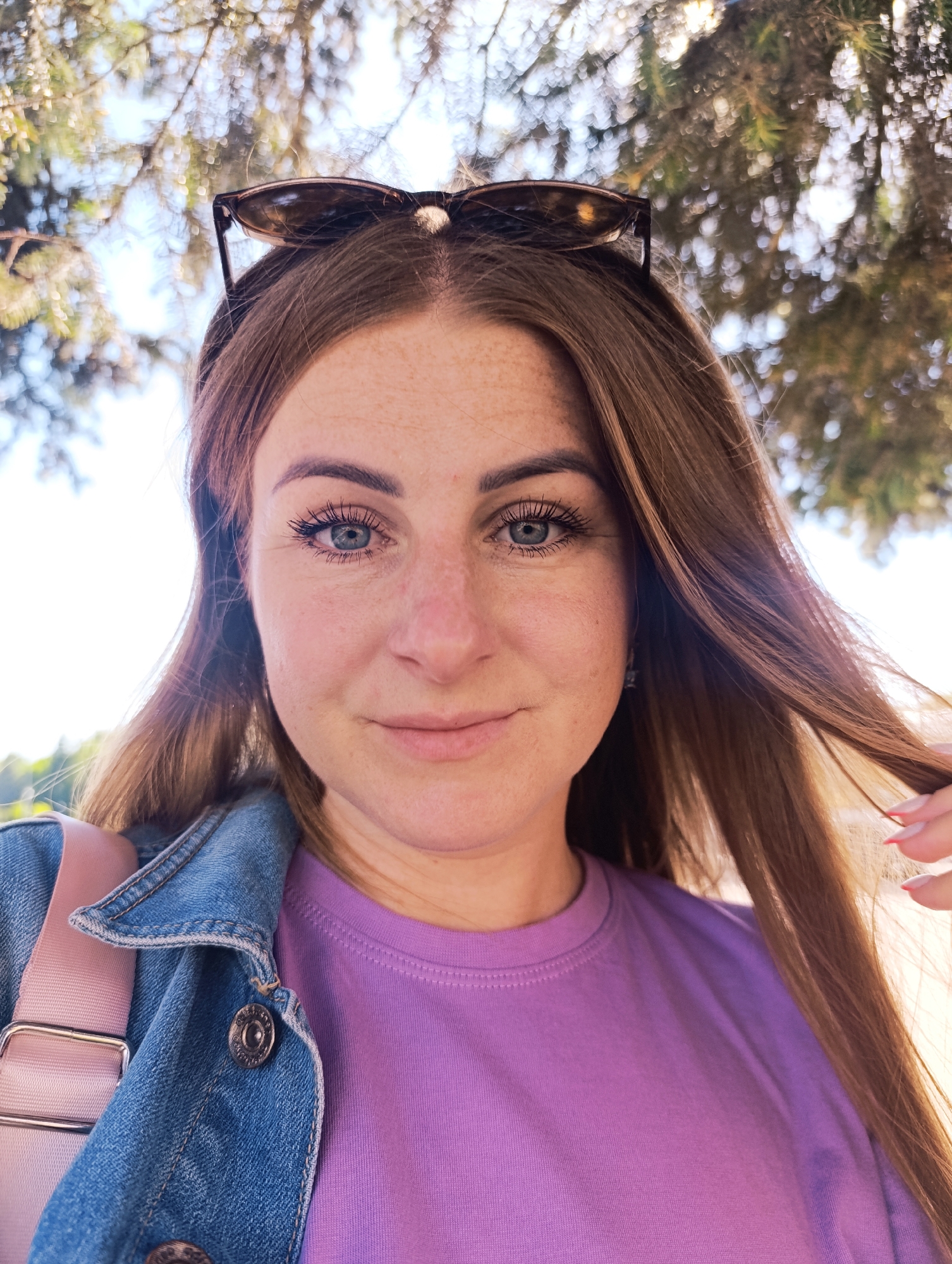 This is a close-up, selfie-style photo of a young woman outdoors, likely during the daytime under a tree. She has long, light brown hair that is styled down and slightly pulled to the side by her right hand. Sunglasses are perched on top of her head. She is wearing a light purple t-shirt and a blue denim jacket, with a pink strap from a bag visible over her shoulder.
The background is blurred, showing dappled sunlight filtering through green foliage, suggesting an outdoor setting such as a park or forest. The lighting indicates it is daytime, likely sunny given the bright highlights. The woman's expression is a gentle smile, and she appears relaxed and happy. She has light skin with freckles and blue eyes, and her makeup includes mascara. No visible text is present in the image.