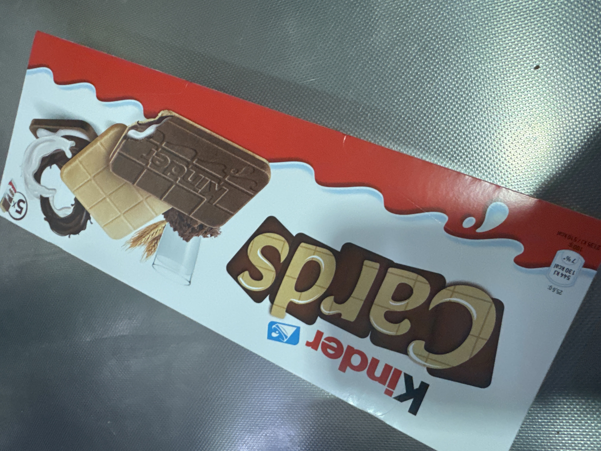 This is a close-up, overhead shot of a Kinder Cards box. The box is white with red and blue accents, and features images of the chocolate product. The word "Kinder" is prominently displayed in red, followed by "Cards" in large, brown, three-dimensional letters. The lighting suggests an indoor setting, possibly a kitchen or grocery store, with the box resting on a textured, metallic surface. The location in Parekklisia Community, Cyprus is not discernible from the image itself.