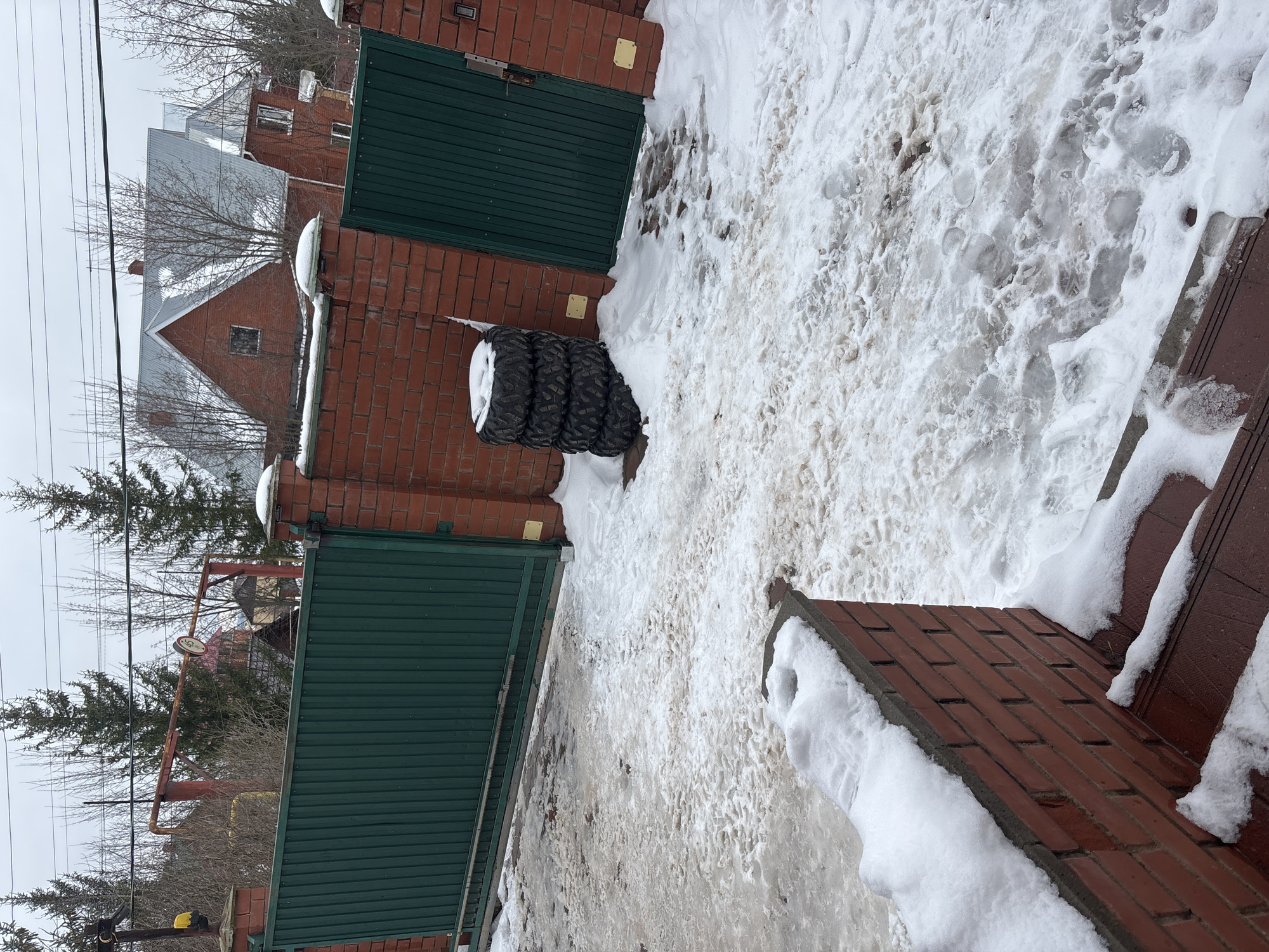 The image depicts an outdoor scene in Ферма, Russia. The ground is covered with a mix of snow and ice, showing signs of melting and footprints. A red brick gate structure with dark green metal gates is visible. A stack of three tires leans against one of the brick pillars. In the background, there are trees, overhead power lines, and rooftops of buildings. The sky is overcast.