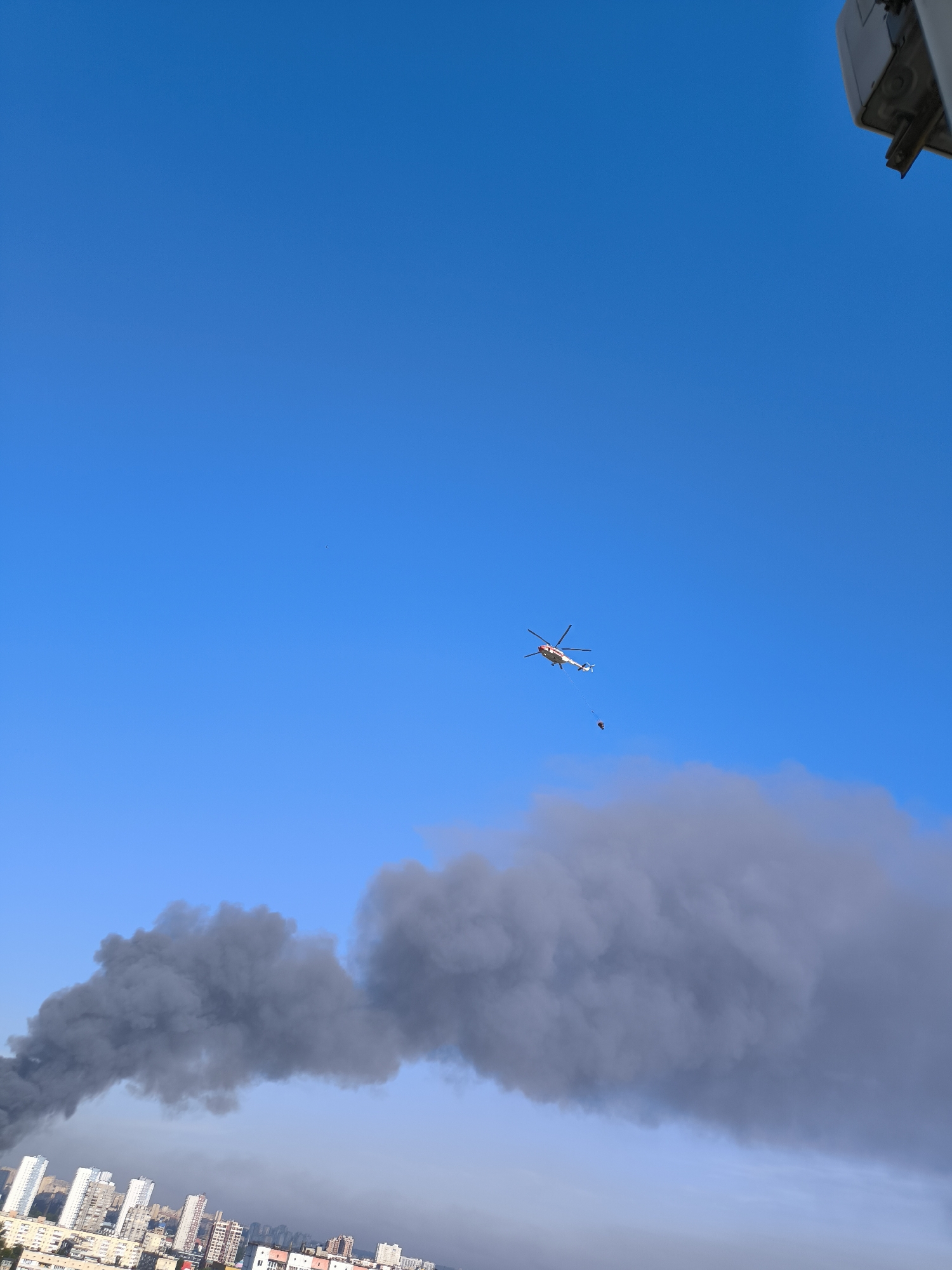 In a sky of clear blue, a white and red helicopter with a suspended bucket is seen flying over a city. Below the helicopter, a large plume of dark grey smoke billows across the sky, obscuring a portion of the city's skyline, which consists of numerous buildings of varying heights. The smoke appears to be emanating from a source within the city, though the exact origin is not visible. The helicopter is positioned above the smoke and seems to be either observing the situation or preparing to assist in extinguishing a fire. The overall scene suggests an emergency response in an urban environment.