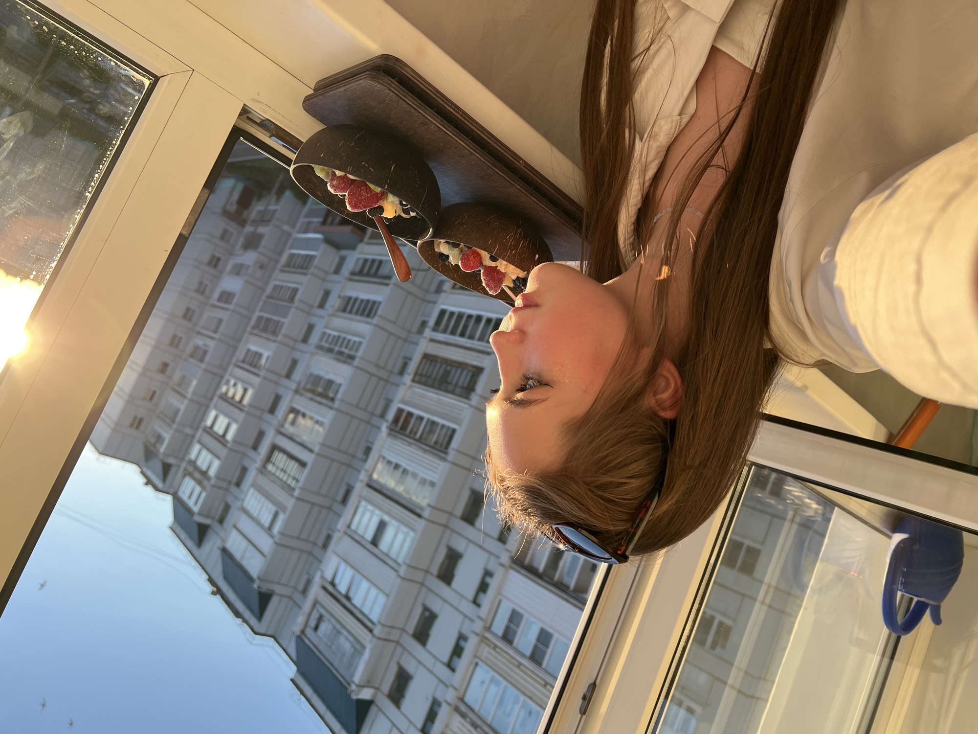 The image, originally oriented on its side, depicts a young person indoors near a window, enjoying a meal while looking out at an urban scene. A person with long, straight brown hair, wearing a light-colored collared shirt and sunglasses pushed onto their head, is seen in profile from the chest up on the right side of the frame. Their gaze is directed upwards and slightly to the right, suggesting a calm and contemplative mood.
In front of the person, on a dark rectangular tray, are two coconut bowls. Each bowl is filled with a colorful assortment of fruit, including strawberries, blueberries, and banana slices, combined with what appears to be yogurt or a smoothie. A small wooden spoon rests in one of the bowls.
The background shows a large window, which has some streaks or dirt on the glass. Through the window, a bright, clear sky is visible, along with the facade of a multi-story, light-colored apartment building featuring numerous rectangular windows and balconies. The architectural style suggests an urban environment typical of Eastern Europe or post-Soviet regions, consistent with Chaikovsky, Russia. The strong, warm light on the left side of the window and reflecting on the person's hair and face indicates late afternoon or early evening, likely during the golden hour or sunset. A blue mug is partially visible, reflected in the window in the bottom right corner. No text is visible in the image.