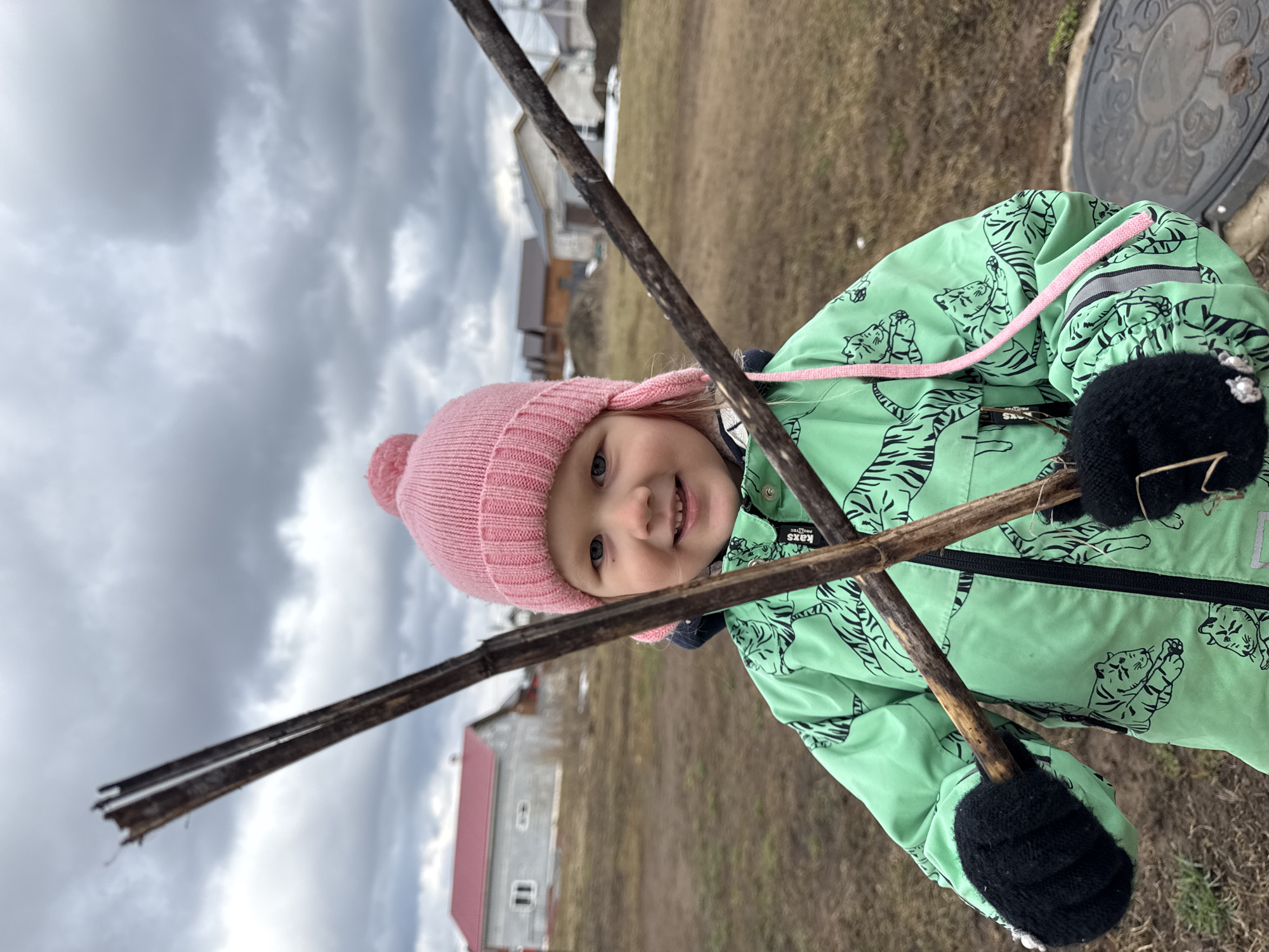 In Gornyi (Dvurechenskoe S/P), Russia, a young child wearing a pink knit hat and a light green tiger-striped jacket stands outdoors, holding two sticks in front of them. The child is also wearing black mittens. In the background is a landscape with a cloudy sky, patches of snow, and a few buildings. A manhole cover is visible to the right of the child.