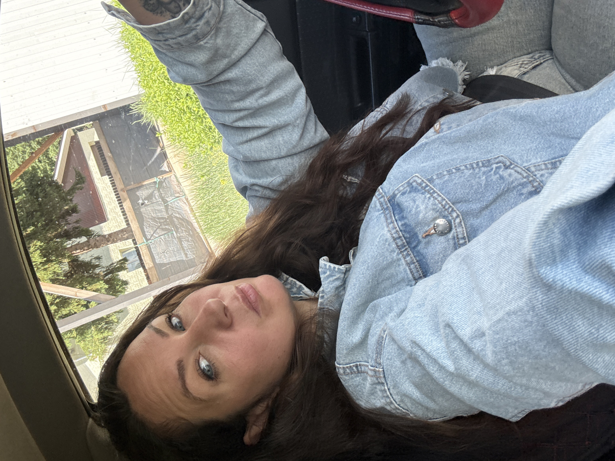 A woman is lying in a vehicle, possibly the passenger seat, with her head near the window. She has long brown hair and blue eyes, and she is wearing a denim jacket and denim jeans. The vehicle appears to be parked or stationary. Through the car window, a building is visible, possibly a house, and trees are seen. The building features a brown exterior. The location may be in Chaikovsky, Russia.