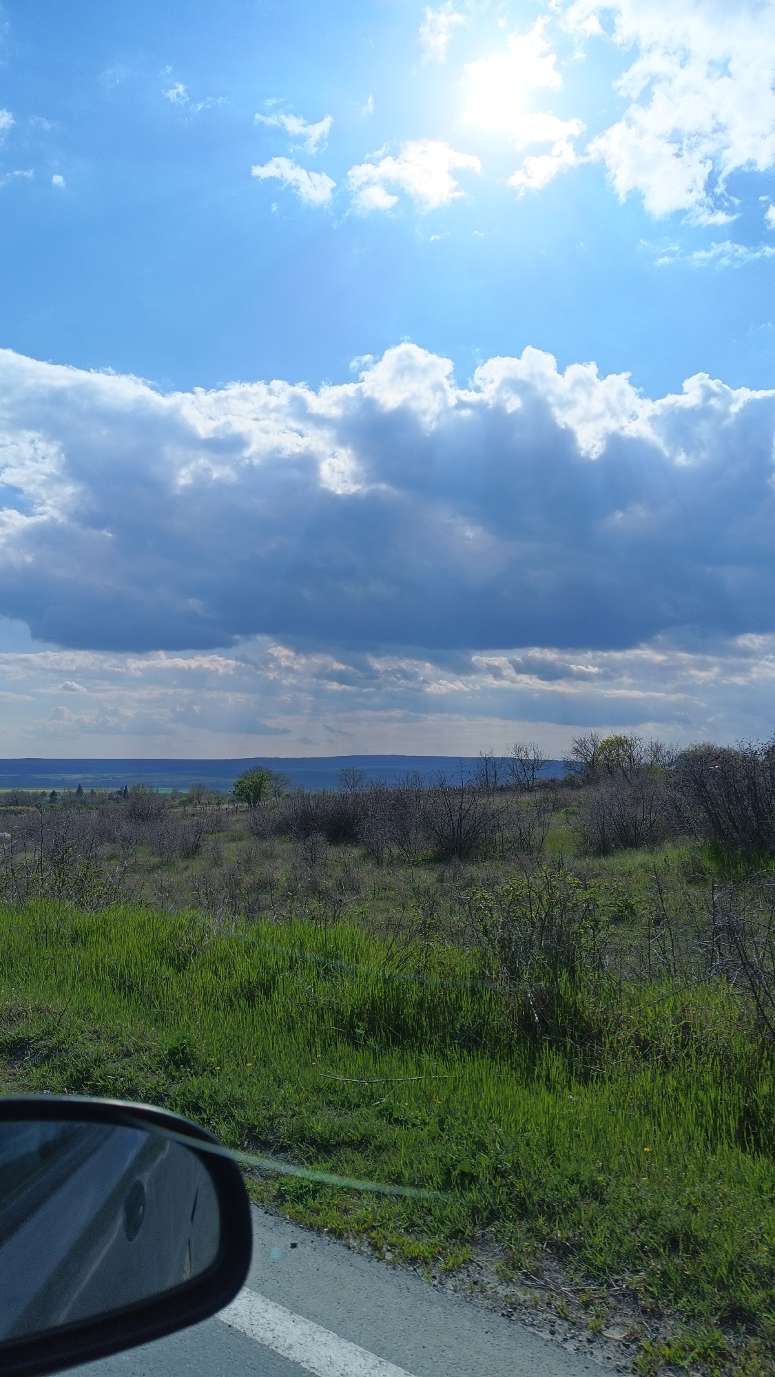 The image depicts an outdoor scene on a sunny day with clouds, likely taken from a moving vehicle. The foreground includes a section of paved road with a white painted line and a side mirror of a car is partially visible. Beyond the road, there is an expanse of grassy terrain. In the middle ground, there are shrubs. The background consists of a slightly elevated landscape, and beyond that a more distant and hazy view of what could be a body of water or other terrain. The location is likely near Valchin, Bulgaria. There are no people visible in the image.
