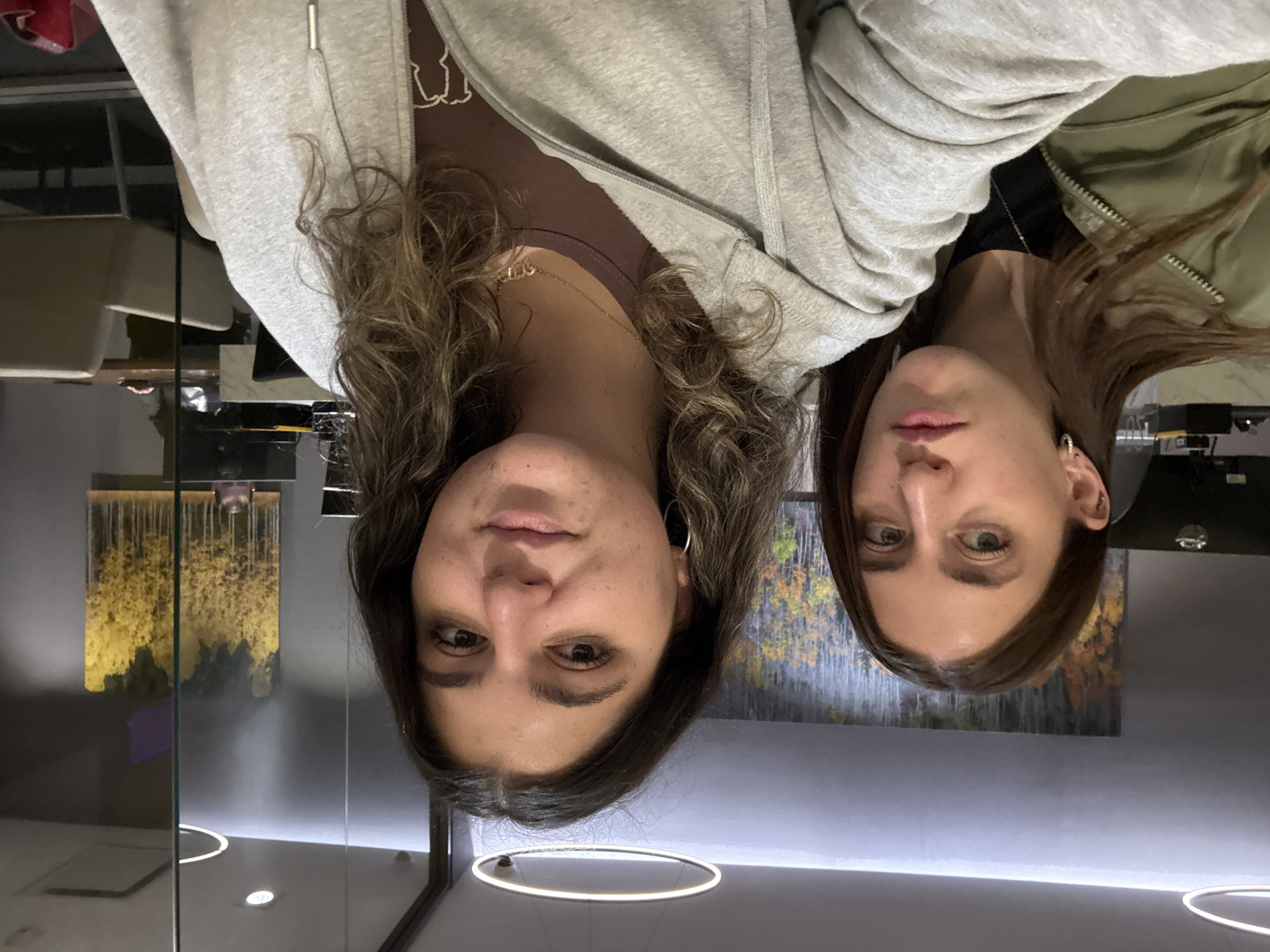 This image depicts two young adult females in a modern, indoor commercial or office setting in Mexico City. The subject in the foreground, on the right, has medium-length wavy brown hair, dark eyes, and is wearing a light grey zippered hoodie over a brown t-shirt with a gold-colored necklace and silver hoop earrings. She exhibits a neutral expression with a slight smile and is looking directly at the camera. The second subject, positioned slightly behind and to the left, has straight brown hair, light-colored eyes, and is wearing an olive green zippered jacket over a dark shirt. She has small hoop earrings in both ears and a helix piercing in her left ear. Her expression is neutral, with slightly pursed lips, and she is also looking at the camera.
The background features dark grey walls and glass partitions. On the wall to the left, a large painting displays abstract vertical forms resembling golden and dark trees. Overhead, a circular, illuminated light fixture is visible on the ceiling. To the right, through and reflected in a glass partition, another large painting with similar golden tree-like forms can be seen. Beyond the glass, the top of a male figure's head is partially visible behind a desk or workstation. Further into the background, beige office chairs and a table are discernible, suggesting a meeting or work area. Another circular ceiling light fixture is reflected in the glass. A red object, possibly a bag, is partially visible on the floor in the bottom right corner of the frame. The environment indicates a contemporary commercial interior, typical of business districts within Mexico City, such as Polanco, Reforma, or Santa Fe.