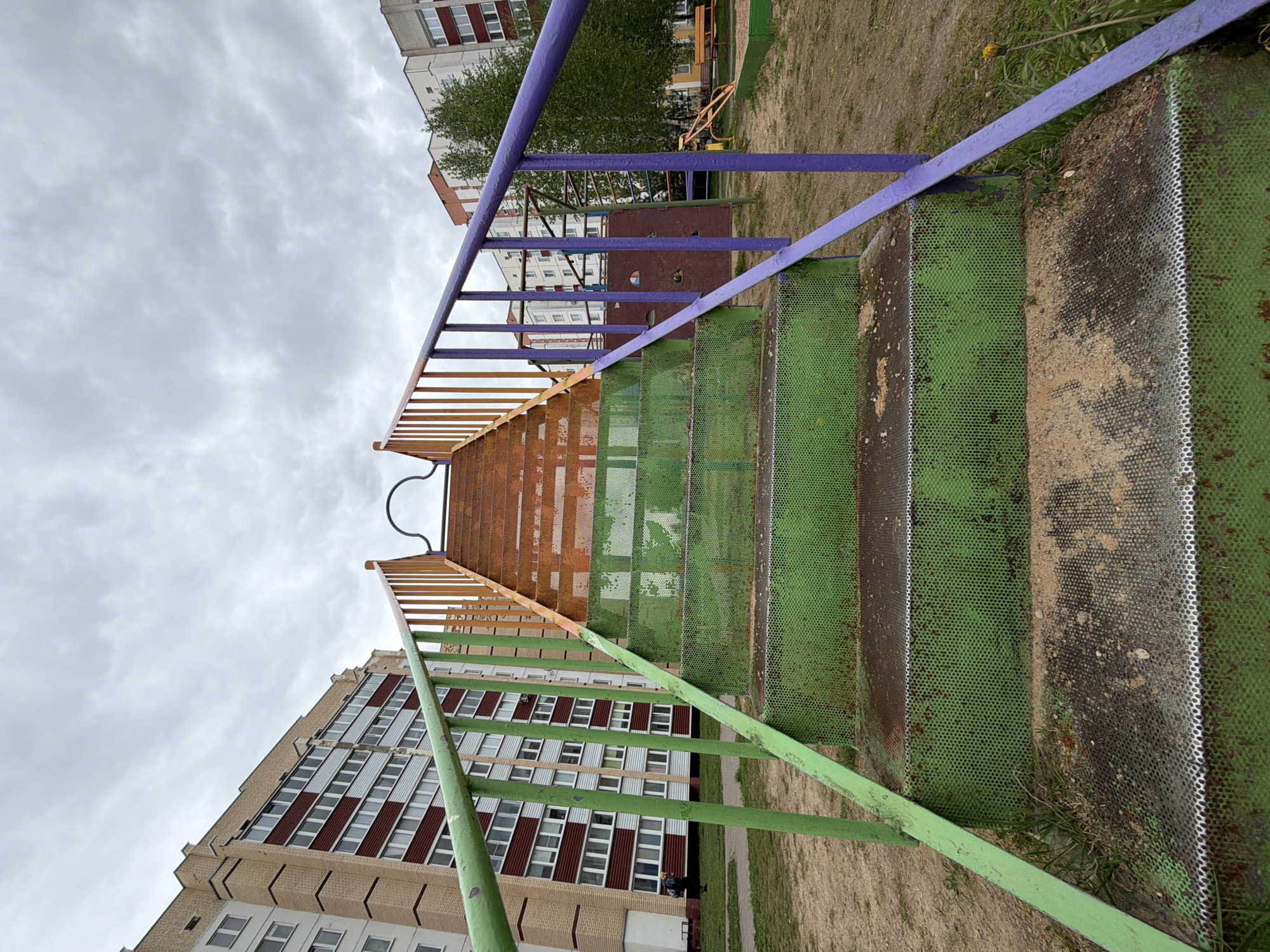 A playground slide dominates the frame, featuring green perforated steps leading to a brown metal platform, with green, purple, and yellow railings. Adjacent to the slide, a tall building with a beige and maroon facade is visible, its windows reflecting the overcast sky. The sky is filled with gray clouds. The ground surrounding the slide is covered in sand and sparse grass. In the background, additional buildings and playground equipment can be seen. The location is a playground in the city of Unknown, Unknown.
