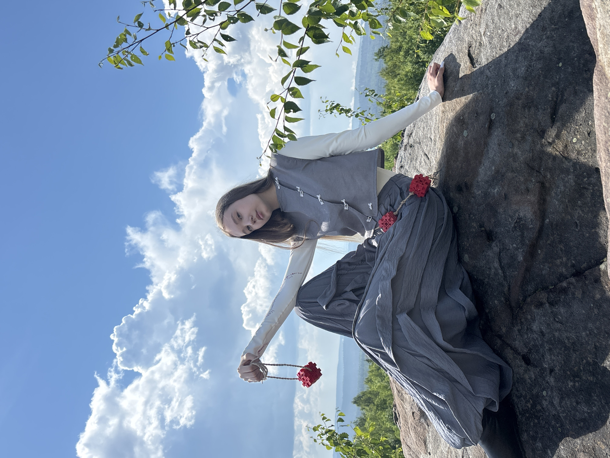 A young female is sitting on a large rock on a sunny day. She is wearing a gray vest over a white long-sleeved shirt and a long gray pleated skirt. She is holding a red cube-shaped pendant on a string in her right hand. The background shows a landscape of green trees and hills under a blue sky with white clouds. The location is Krivel's Mountain in Gubakha, Russia.