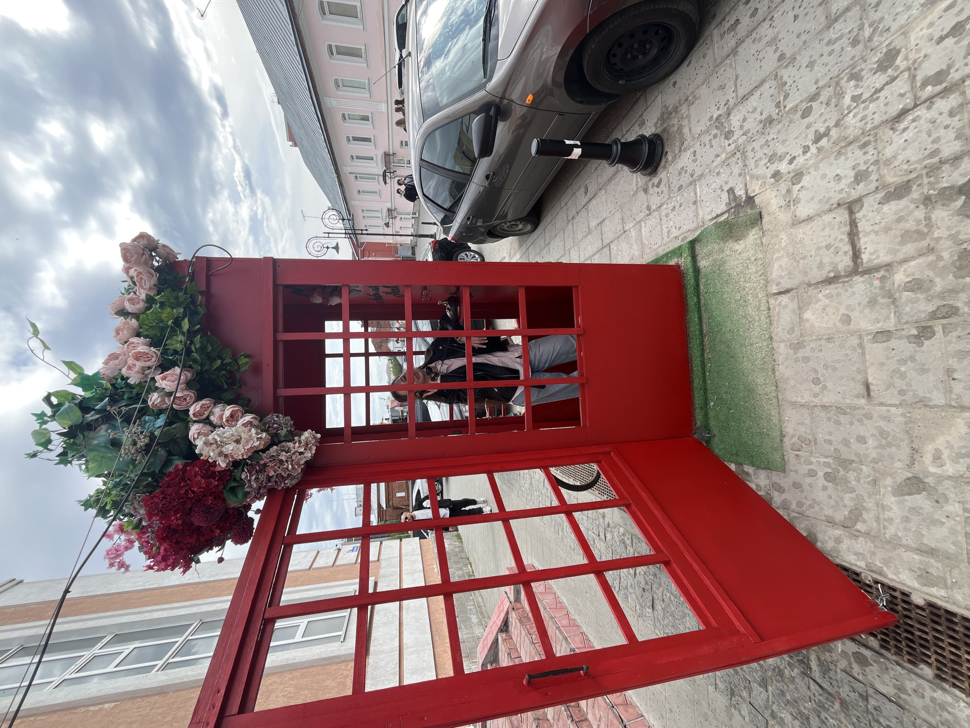 A red telephone booth stands open on a paved street in Ulyanovsk, Russia. The booth is decorated with flowers, including pink roses and burgundy blooms. A person wearing sunglasses, a black jacket, and light-colored pants is sitting inside the booth. A gray car is parked nearby, adjacent to a short, black and white bollard. The facade of a pink building with multiple windows is visible in the background. The sky is cloudy. A patch of green material is placed on the ground next to the telephone booth. A metal grate drain is visible on the right side of the frame.