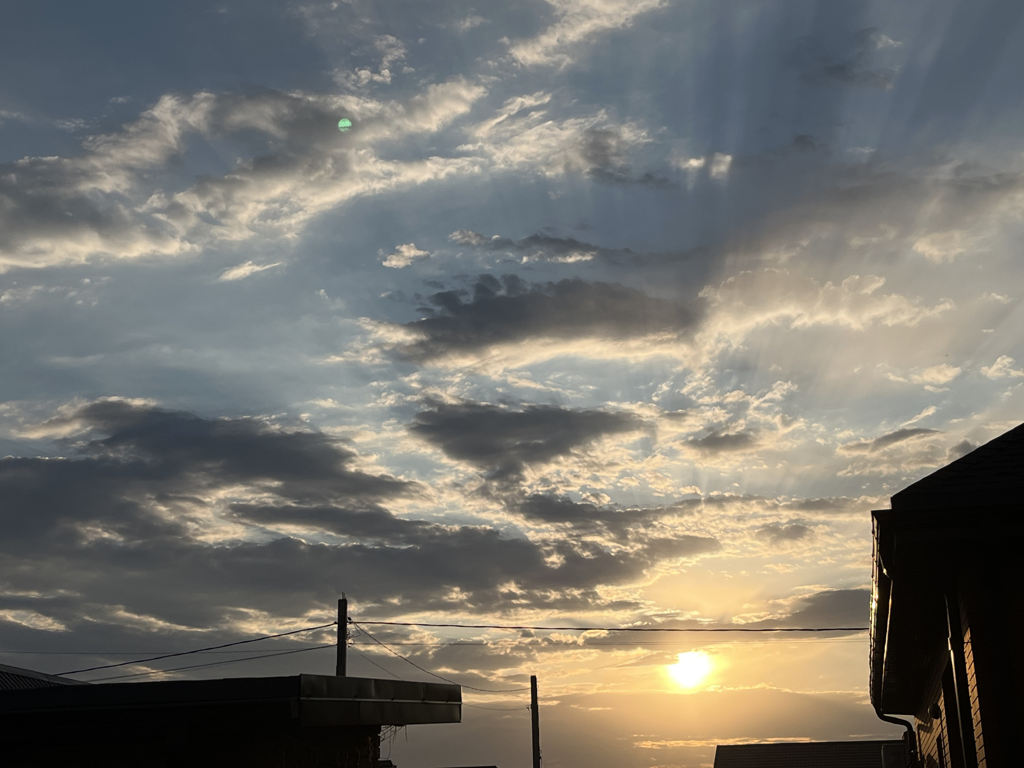 The image depicts a cloudy sky with dramatic sunbeams breaking through the clouds at sunset. The sun is low on the horizon, casting a warm glow. Silhouetted buildings and utility poles are visible in the foreground, suggesting an urban or suburban environment. One of the buildings in the foreground appears to be a house with a dark roof. The sky is a mix of blue, gray, and yellow. A small, bright green circular anomaly is present in the upper left quadrant of the image, which may be a lens flare or a photographic artifact. The scene is likely captured in Taganrog City District, Russia, given the context.