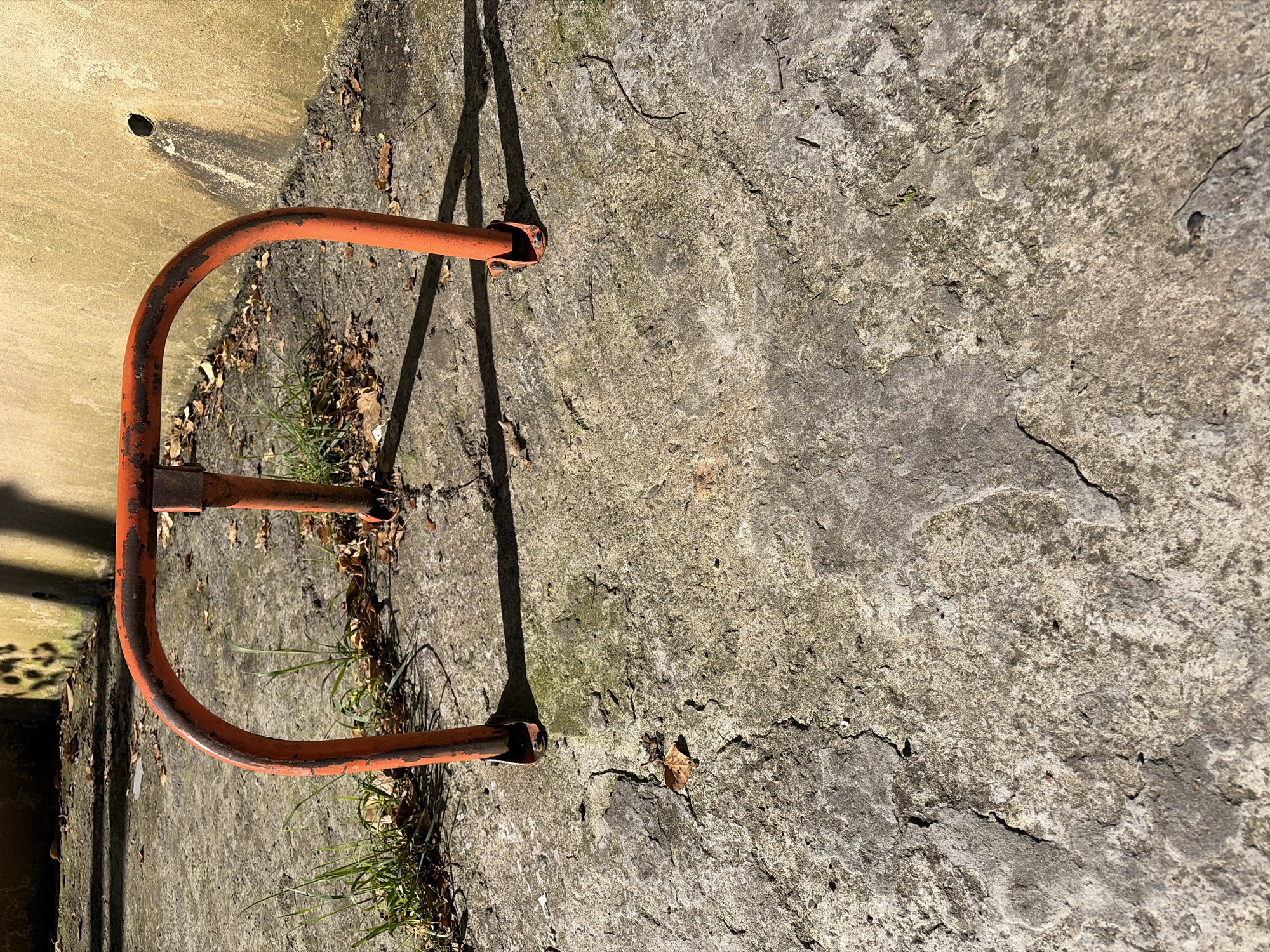 An outdoor scene captures a rusty, U-shaped orange metal barrier anchored to a weathered concrete ground. The barrier, showing signs of paint peeling and corrosion, stands against a textured, yellowish-beige wall on its left. Bright sunlight casts long, distinct shadows of the barrier and potentially other unseen objects across the ground, indicating either early morning or late afternoon.
The concrete surface is rough, cracked, and covered with patches of moss or dirt. Small clusters of green grass and scattered dry leaves are visible around the base of the barrier and on the ground. No people or activities are present. The overall impression is one of neglect or age, with worn materials dominating the composition. There is no visible text.