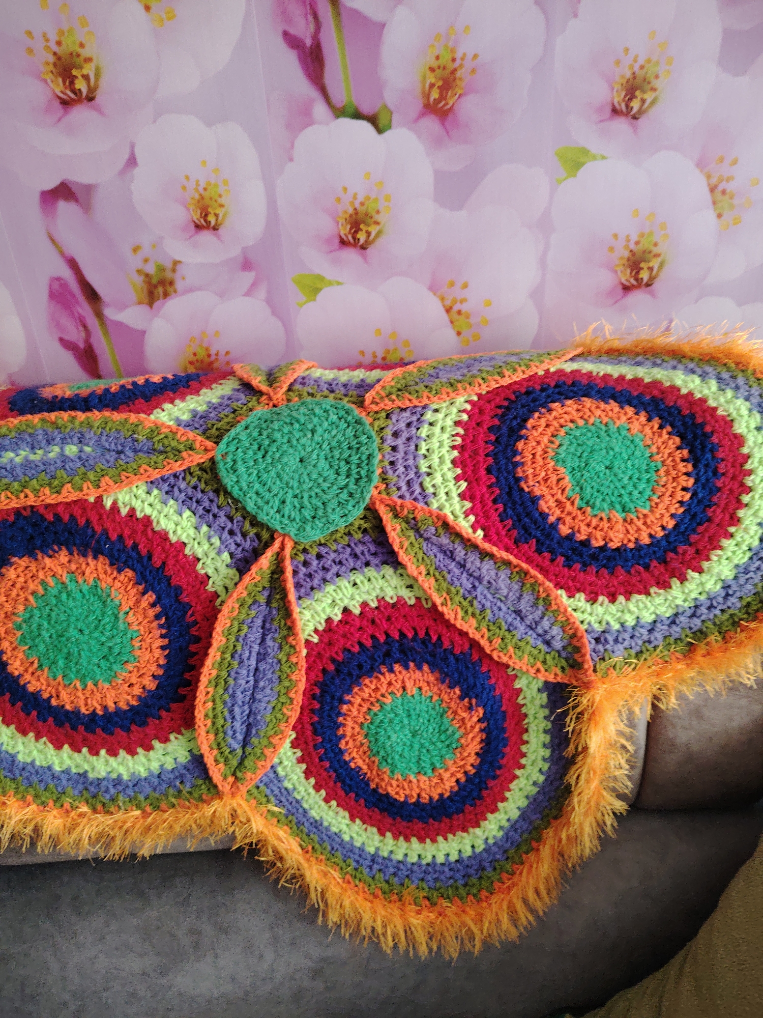 A vibrant and colorful crocheted item, likely a decorative cushion or blanket, rests on a dark gray sofa in an indoor setting. The crocheted piece features a striking design composed of multiple concentric circles, primarily in shades of green, orange, red, dark blue, light green, and purple. A distinct bright green heart-shaped element is centrally placed on the item, with two smaller, leaf-like crocheted extensions branching out from it. The entire perimeter of the crocheted piece is edged with a fluffy, bright orange yarn, adding a soft, textured border.
Behind the crocheted item, a backdrop or wall covering displays a pattern of light pink and white cherry blossoms (sakura) with bright yellow stamens, creating a soft, floral aesthetic. The lighting suggests daytime, enhancing the vividness of the colors. No people are visible, nor is any text present in the image. The scene conveys a sense of handmade warmth and cheerful decor. This image was taken in Yanaul, Russia.