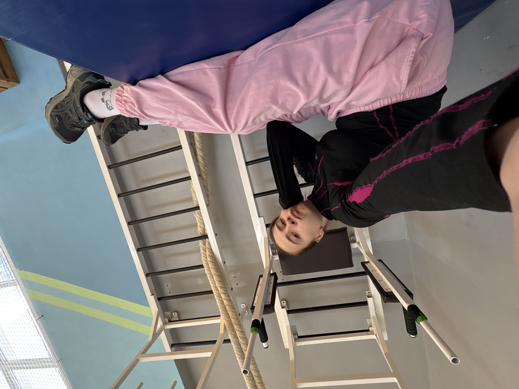 An indoor, vertically oriented shot captures a person performing an inversion exercise, hanging upside down from a Swedish ladder. The individual, appearing to be a young person, is centered in the frame, looking directly at the camera with a calm expression.
They are dressed in light pink, possibly velour, sweatpants or track pants, and a black long-sleeved athletic top with bright pink stitching or accents. Their feet are hooked over a dark blue mat or cushion positioned on the top of the Swedish ladder. They wear dark athletic shoes with light soles and white ankle socks, one of which features a small, light-colored graphic resembling Hello Kitty.
The background reveals a gymnasium setting. A light blue wall with two light green horizontal stripes is visible, suggesting a vibrant and active environment. To the right, a sturdy wooden Swedish ladder with numerous horizontal rungs dominates, with a thick, coiled rope hanging beside it. A removable pull-up bar with green and black grips is attached to the ladder structure. Part of a window with a mesh screen can be seen on the left. The overall lighting appears to be bright and even, consistent with an indoor facility. No explicit text is visible within the image.
The scene takes place in Chaikovsky, Russia.