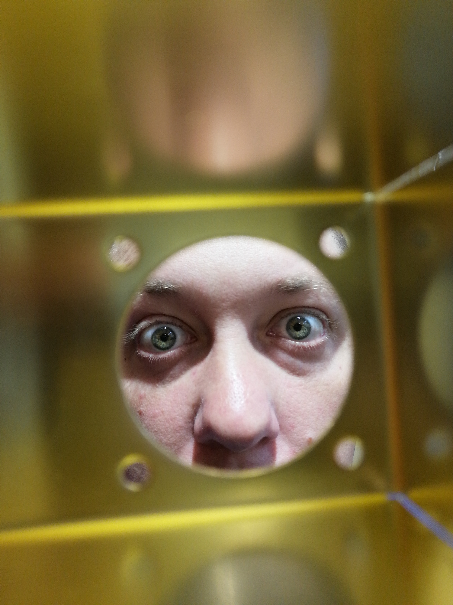 A close-up shot captures a person's face, specifically their eyes and nose, peering through a circular hole in a yellow, possibly metallic, surface. The surface has several other smaller holes around the main one. The person's skin is fair, and their eyes are light-colored. The background is blurry, suggesting a shallow depth of field. There are indistinct shapes and a horizontal line. The location is not discernible from the provided image and cannot be specified as "Unknown, Unknown".