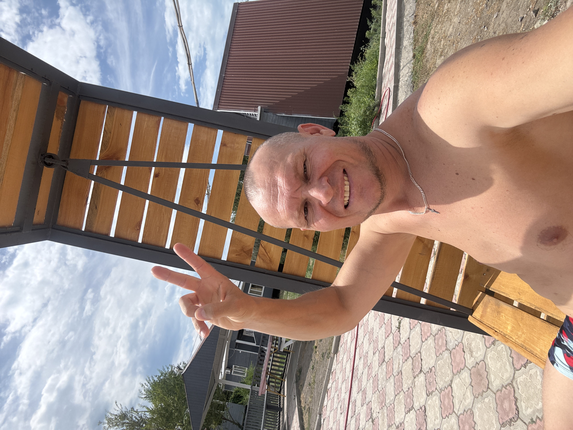 A man is taking a selfie outdoors on a sunny day. He is shirtless and wearing colorful swimming trunks. He is sitting on a wooden bench that is part of a larger structure with wooden slats and a dark metal frame. His left arm is raised, and he is making a peace sign with his fingers. In the background, there are several buildings, including a gray one with white trim and windows, and a brown one with corrugated metal siding. There is also a paved area with a patterned design, green vegetation, and a blue sky with white clouds. The location appears to be a recreational area or a residential compound. The environment suggests a warm climate, possibly a resort or a rural setting within Zharbulak, Kazakhstan.