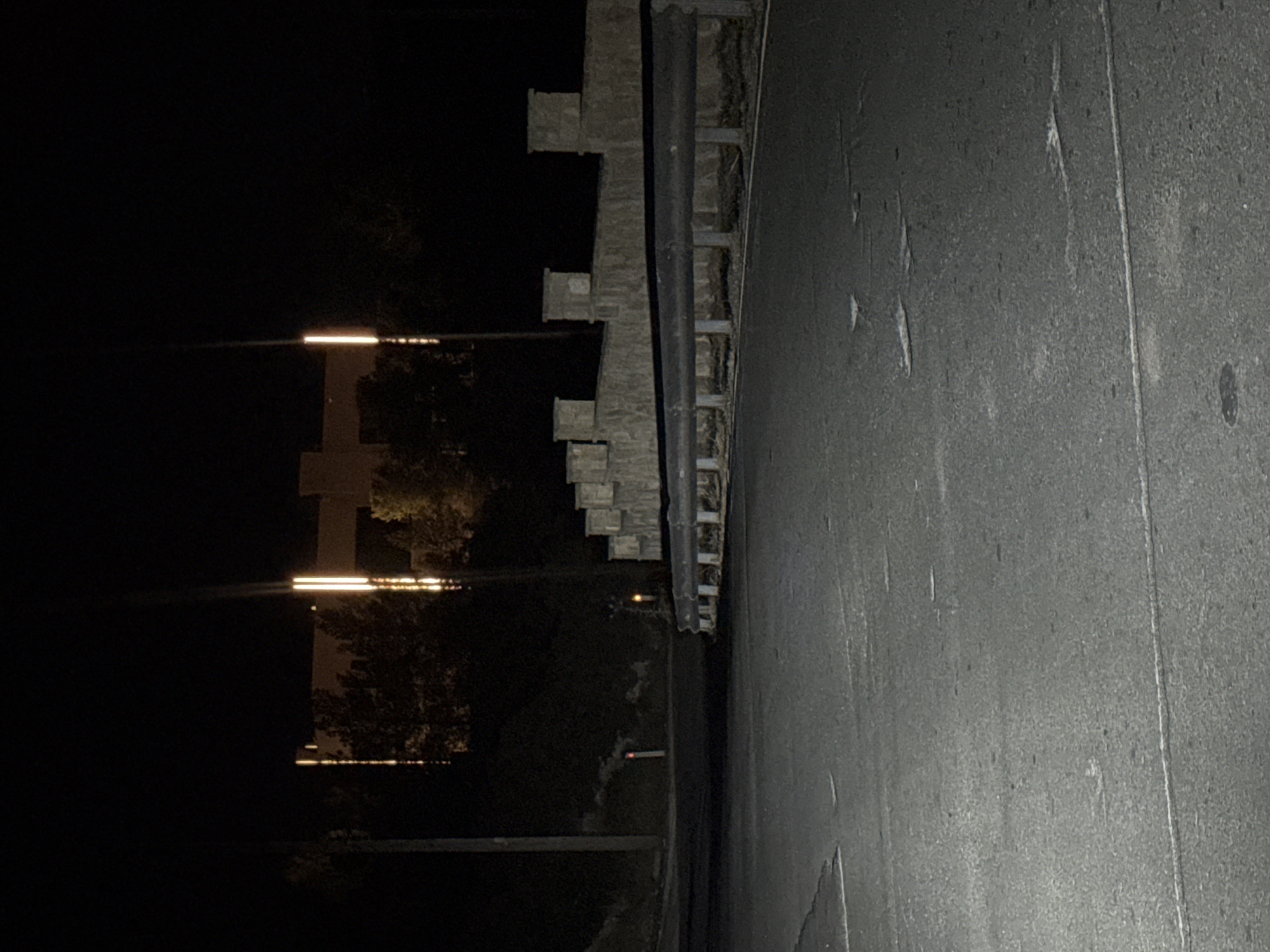 The image captures a dark, nighttime scene on an asphalt road. In the background, a large, illuminated building with several bright vertical lights is visible, surrounded by trees. To the right of the building, a stone wall with decorative pillars runs horizontally across the frame, bordering the road. A metal guardrail is positioned in front of the stone wall. The road surface appears somewhat worn, with visible lines and occasional debris. The overall impression is one of a quiet, possibly rural or semi-urban area at night. The location is identified as Agios Tychona Community, Cyprus.