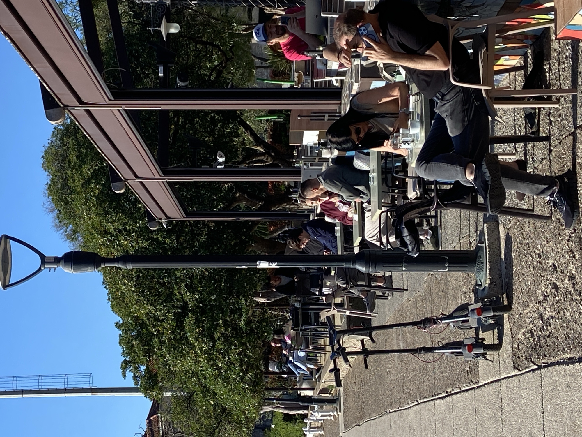 The image shows an outdoor scene in Budva, Montenegro. Several people are seated at tables under a shaded structure, possibly a restaurant or cafe. Some individuals are looking at their phones. Electric scooters are parked along the edge of the sidewalk next to the tables. A black street lamp stands prominently on the left side of the image. Lush trees are visible in the background and along the sidewalk. The sky is clear and blue. The ground appears to be paved with stone or concrete.