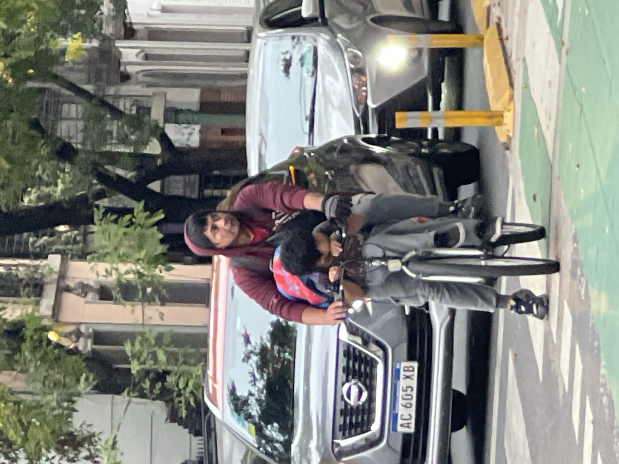 A person is seen riding a bicycle, with a child seated in front. The person is wearing a maroon hooded sweater, a backpack, and gloves. The child is wearing a jacket with a red and blue pattern. They are in front of a parked silver Nissan truck with license plate number AC 605 XB. The background shows urban elements such as buildings and trees. The image was taken in Buenos Aires, Argentina, as evidenced by the license plate of the truck.