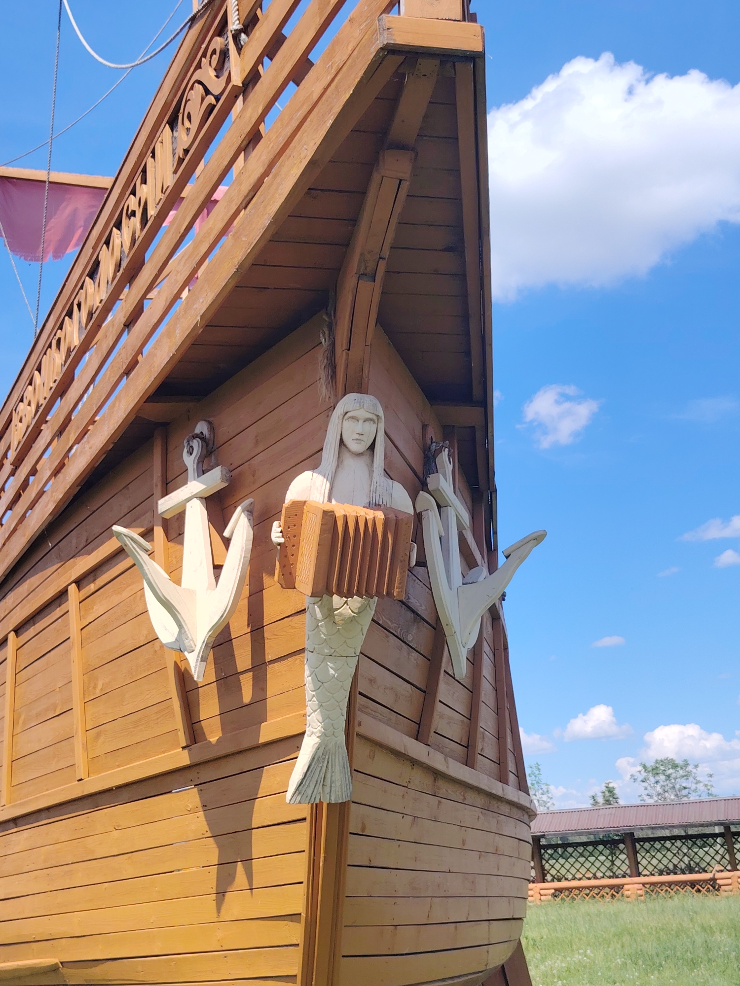 A wooden sculpture of a mermaid holding an accordion is attached to the side of a wooden structure resembling a ship. The mermaid has long, flowing hair and a tail with scales. Two white anchors are mounted on the ship's hull, one on either side of the mermaid. The ship has wooden railings with decorative carvings at the top. A pink flag is visible on the upper deck. The background is a bright blue sky with scattered white clouds. The scene appears to be outdoors, possibly in a park or recreational area, with a grassy field and a wooden fence visible in the distance. The lighting suggests it is daytime, likely sunny.
